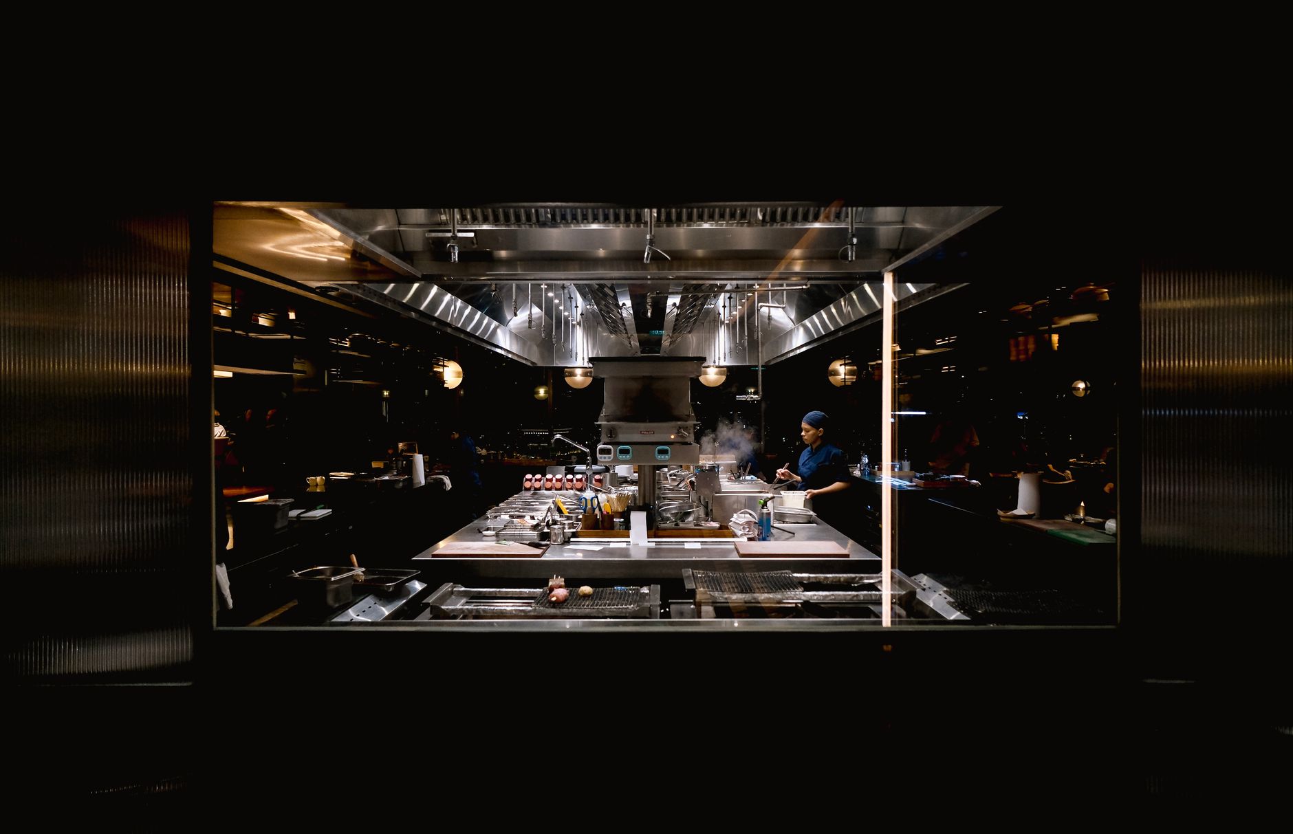 a modern restaurant kitchen with digital order management screens mounted above stainless steel prep stations, late evening service lighting