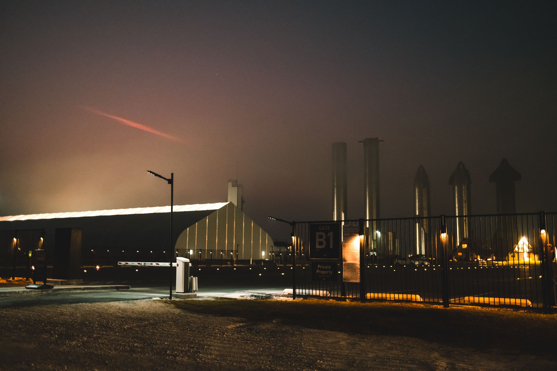 A massive stainless steel rocket booster descending toward a concrete launch mount at dusk, chopstick-style mechanical arms extended outward against a darkening sky