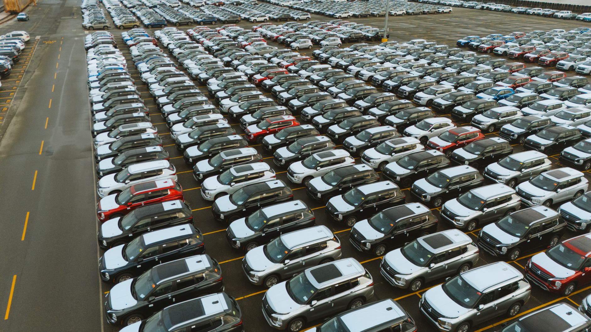 a row of rental cars parked in an airport lot, overhead gantry toll readers visible on a highway entrance ramp in the background at dusk