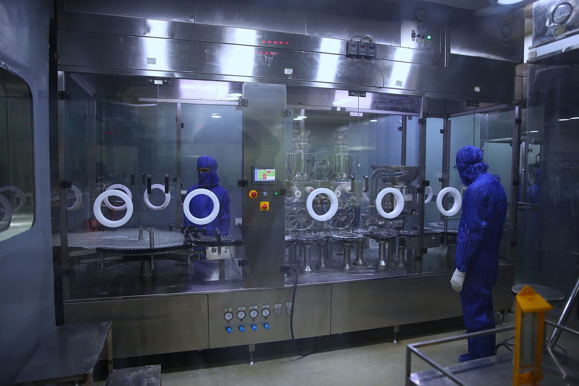 rows of semiconductor wafers arranged in a cleanroom fabrication facility, illuminated by white overhead lighting with robotic handling arms visible in the background