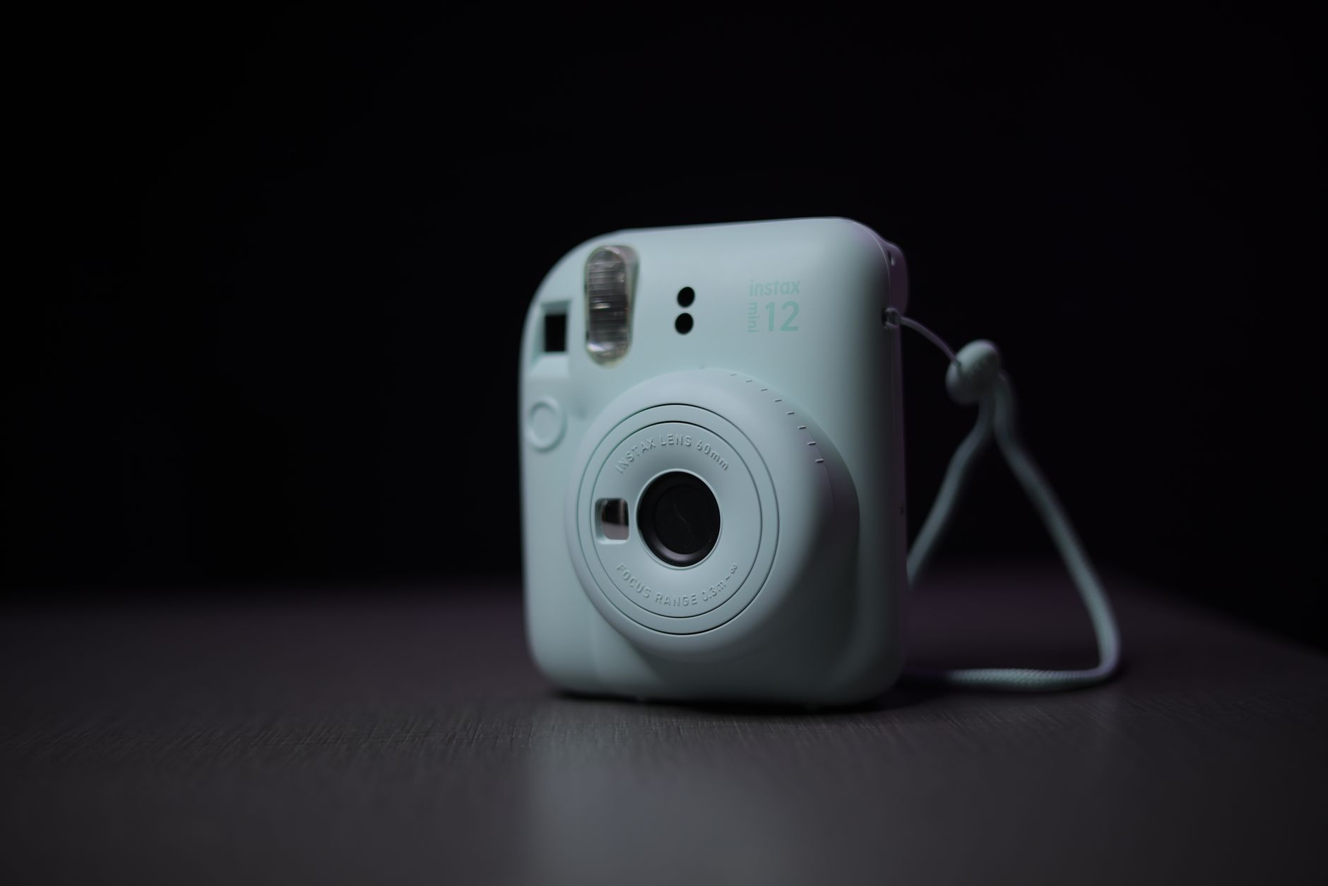 A compact white instant camera resting on a wooden table beside a small stack of square instant photo prints showing soft pastel tones