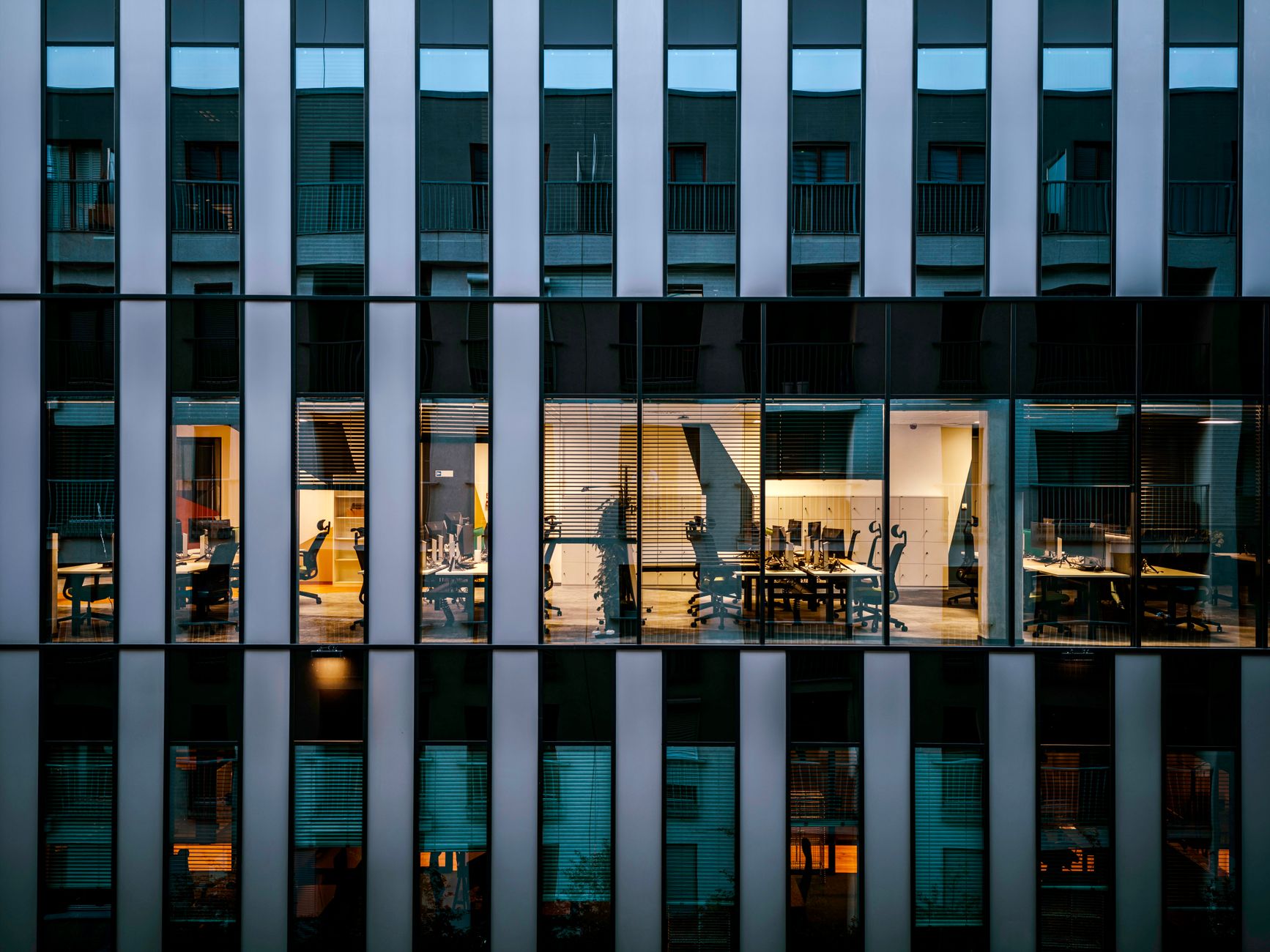a glass-walled modern office building exterior at dusk with interior workstation lights visible through the facade, set against a darkening urban skyline