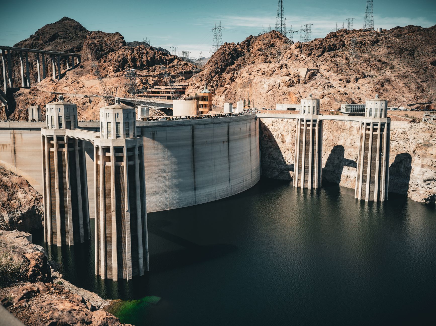 A low-water reservoir in a dry landscape, with exposed sediment and a receded waterline visible along the concrete intake structures of a dam