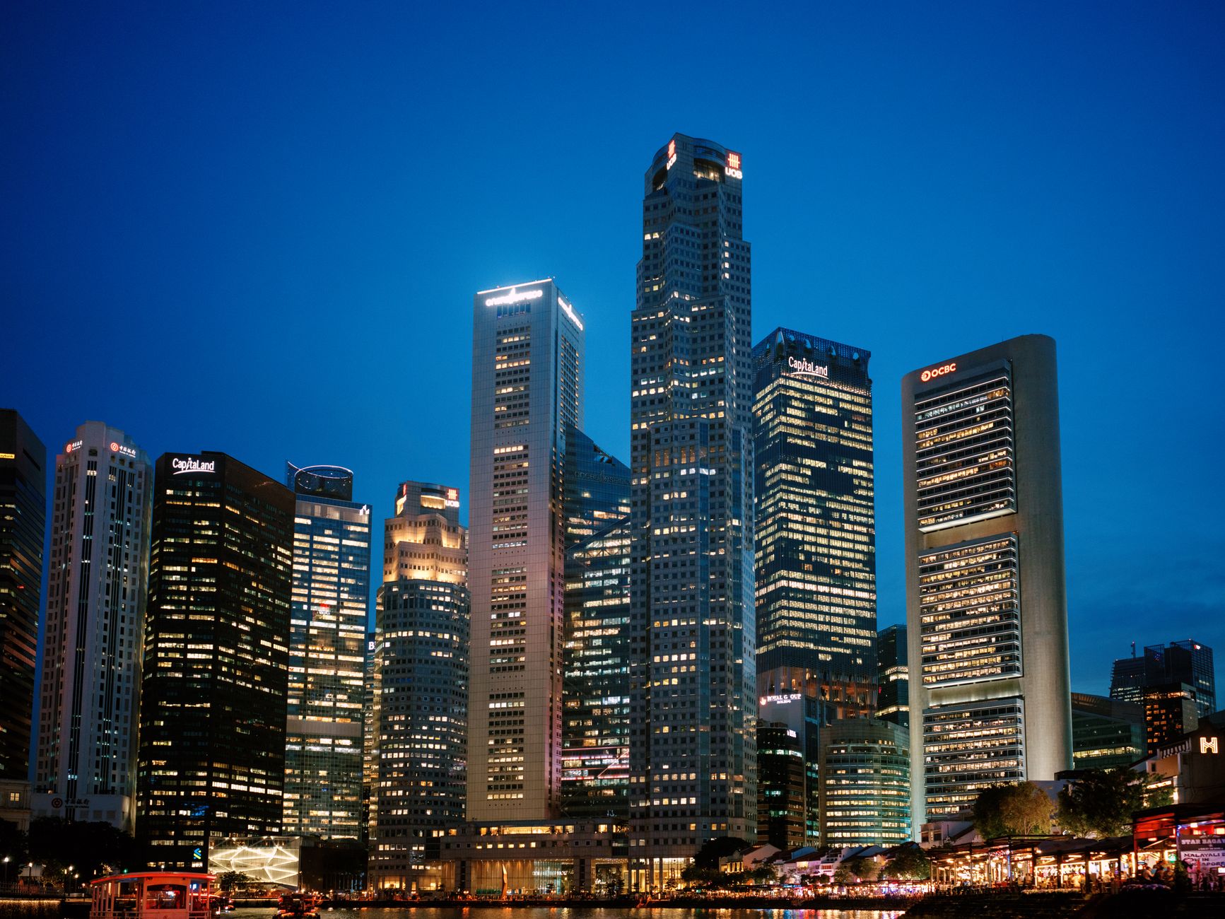 aerial view of Singapore's central business district at dusk, glass towers reflecting orange light, dense urban infrastructure visible below including data center facilities and fiber cable conduits running between buildings