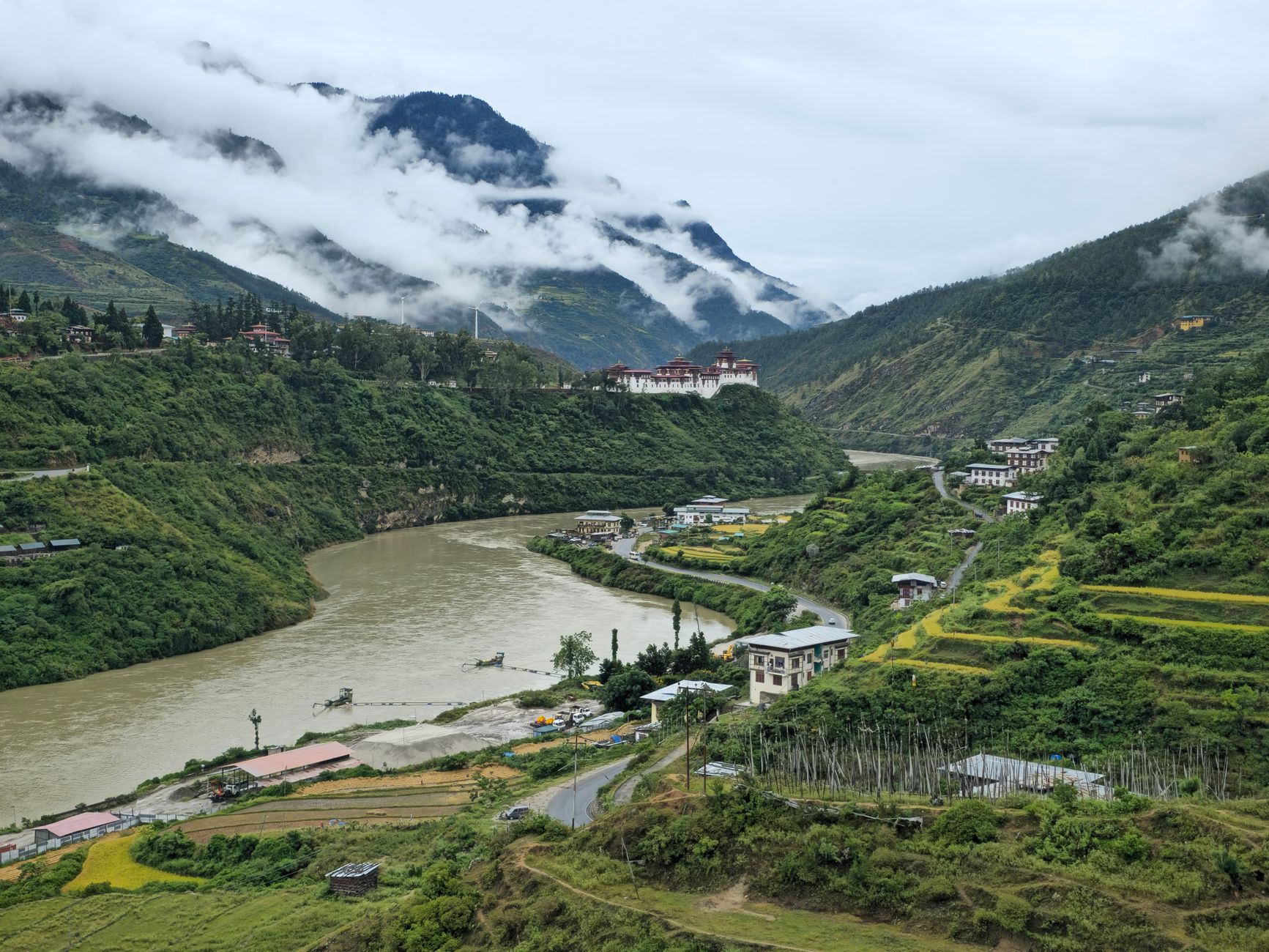 a narrow mountain valley in Bhutan with a cluster of government buildings visible below terraced hillsides, communication towers visible on a ridge in the distance