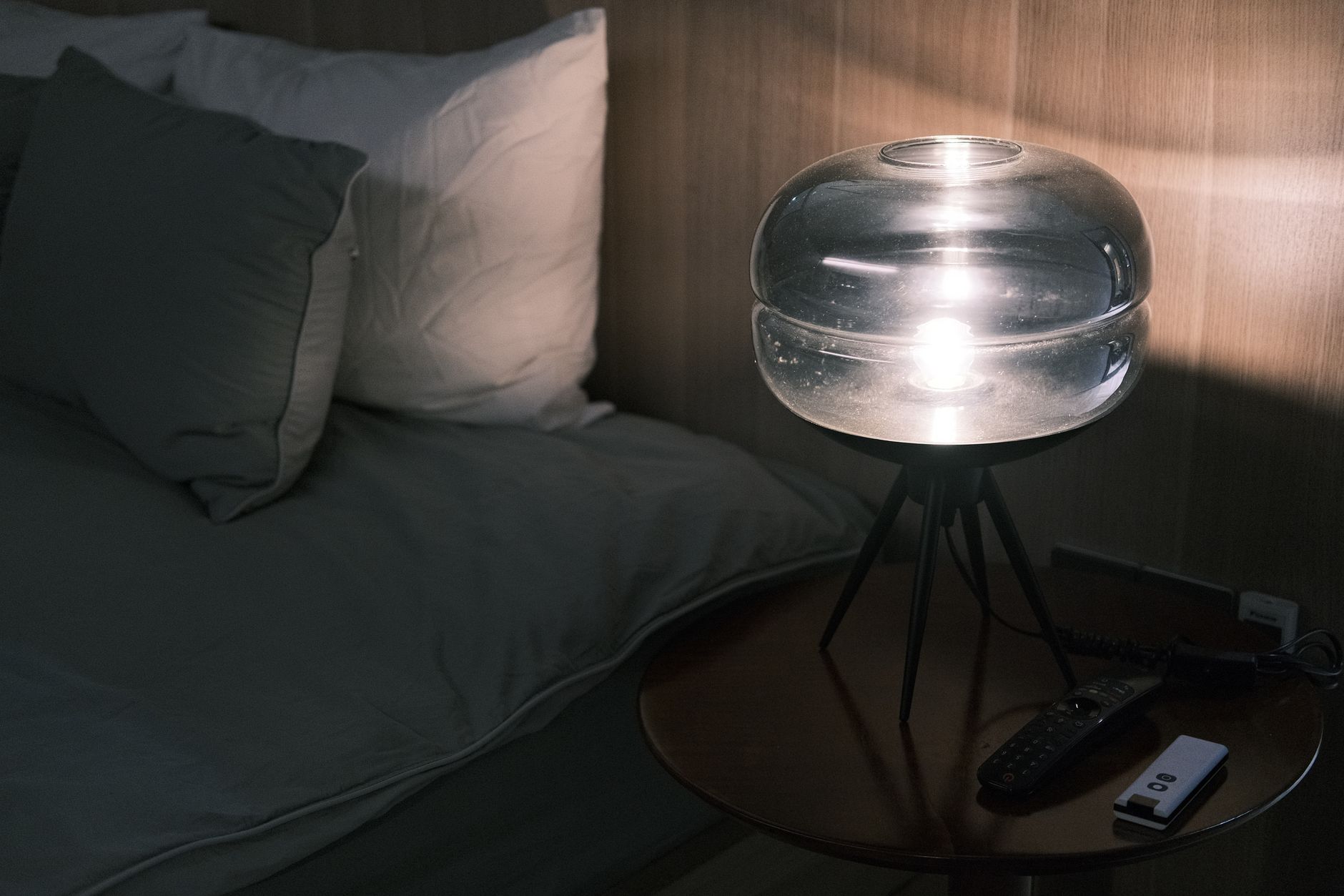 a bedroom nightstand with a dim amber lamp, a glass of water, blackout curtains fully drawn, and a digital clock displaying 1:05 a.m.
