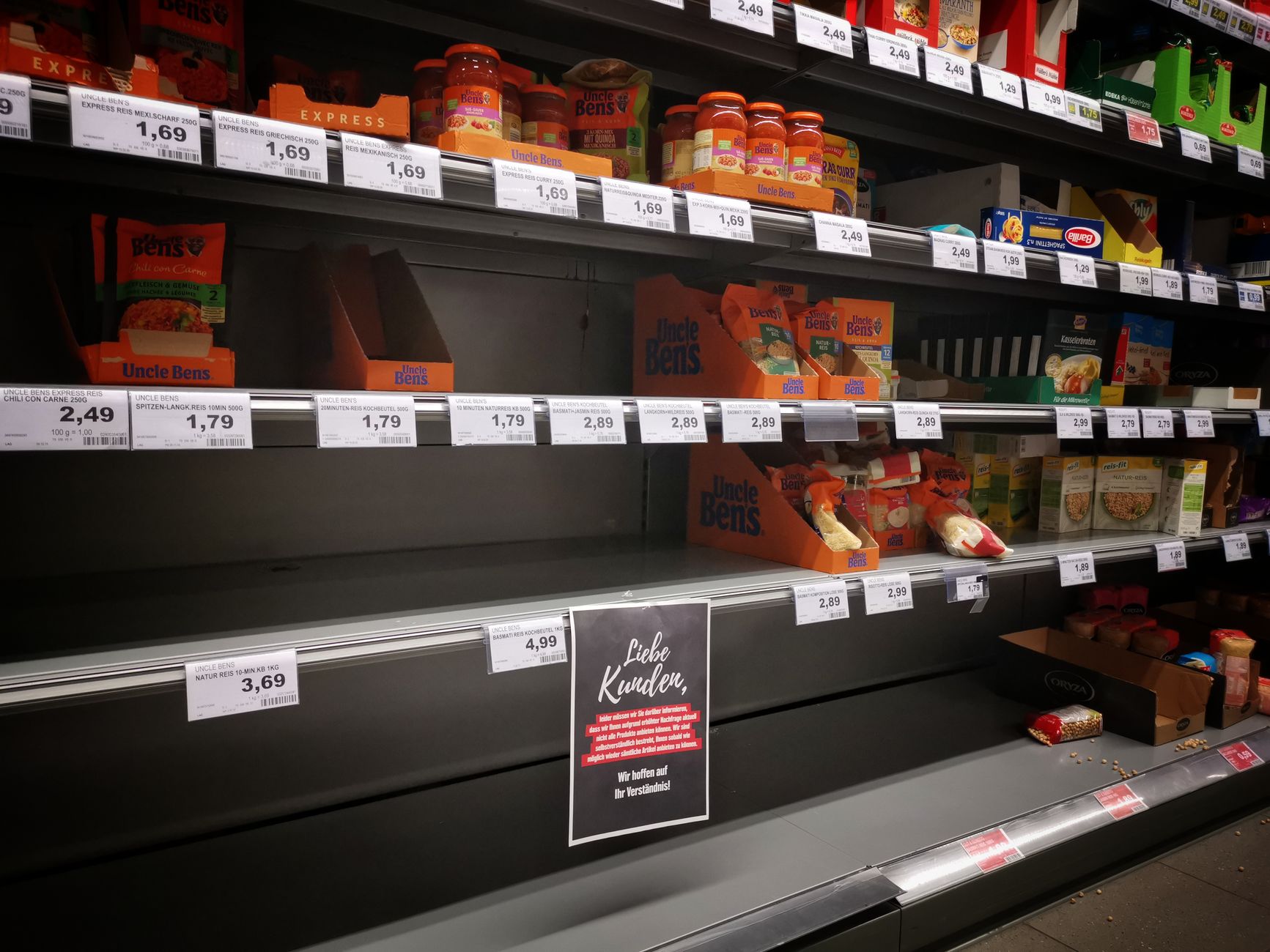 empty shelves of a mid-sized urban grocery store aisle, fluorescent lighting overhead, price tags visible on metal shelf rails