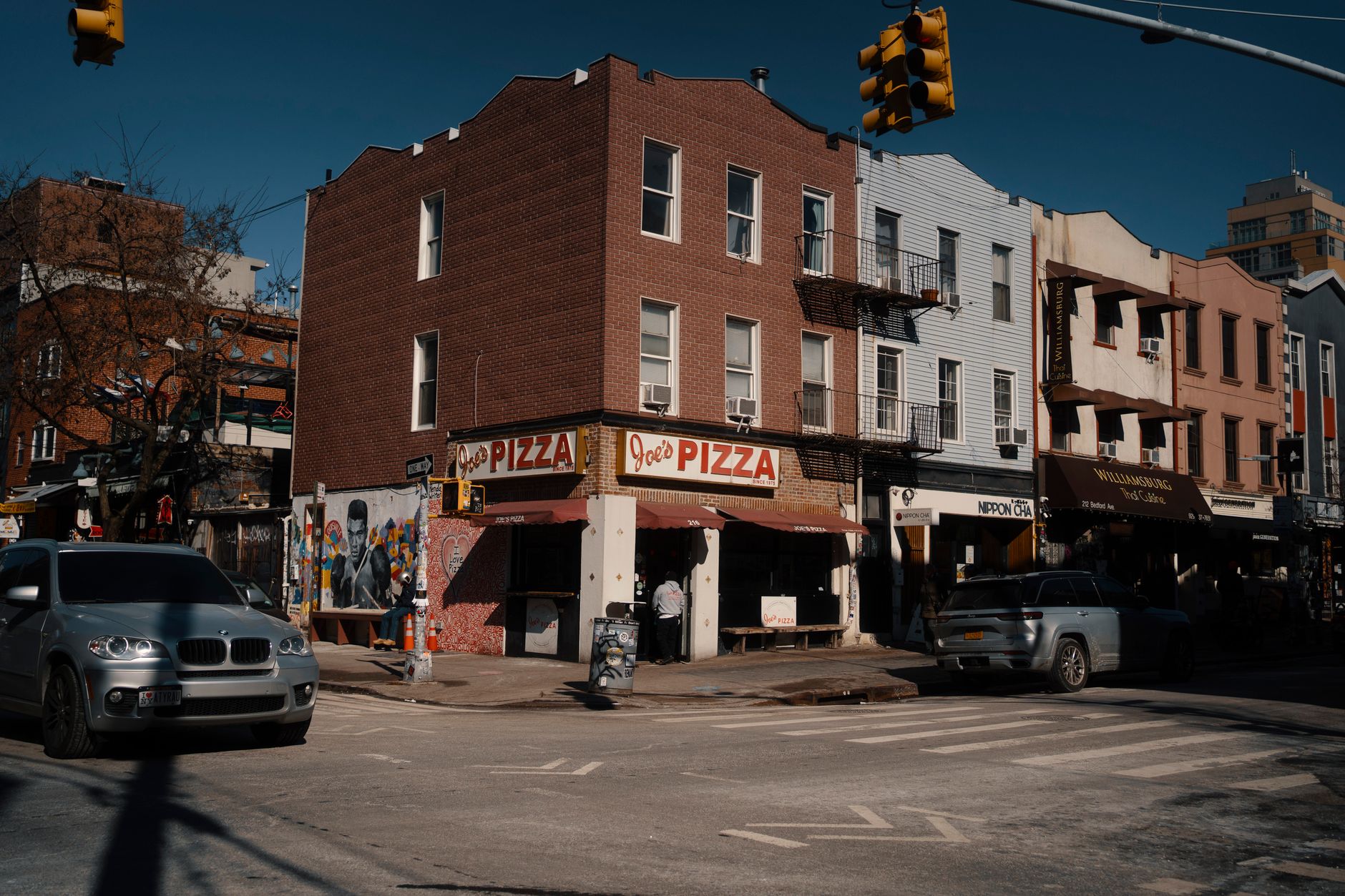 exterior of a corner grocery storefront in a dense urban neighborhood, awning partially visible, sidewalk and parked cars in foreground