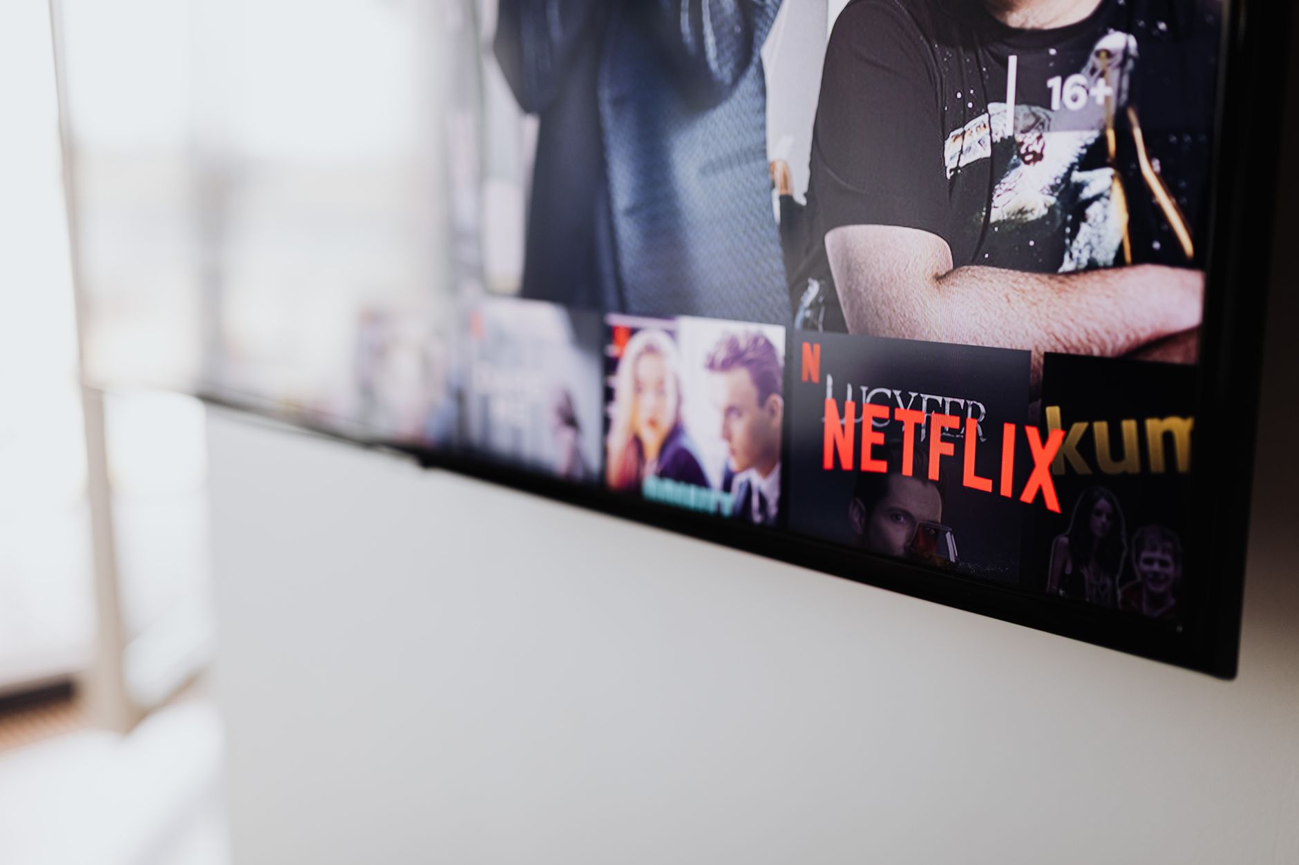a flatscreen television mounted on a living room wall displaying a streaming service home screen interface, with a dark ambient room and a remote control resting on a couch armrest in the foreground