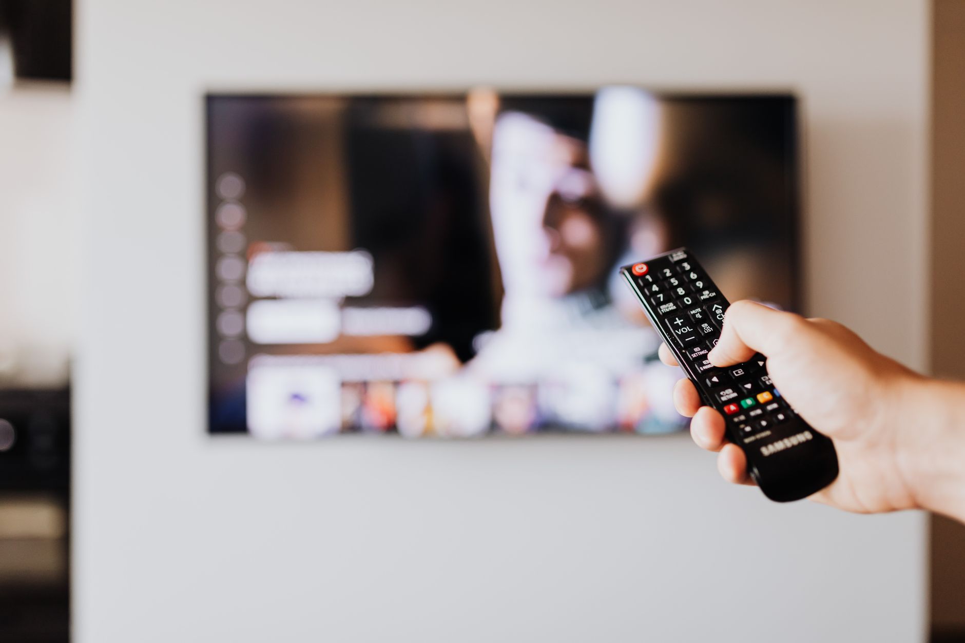 a close-up of a smart television remote control resting on a wooden surface next to a small potted plant, with a softly blurred streaming interface visible on a screen in the background