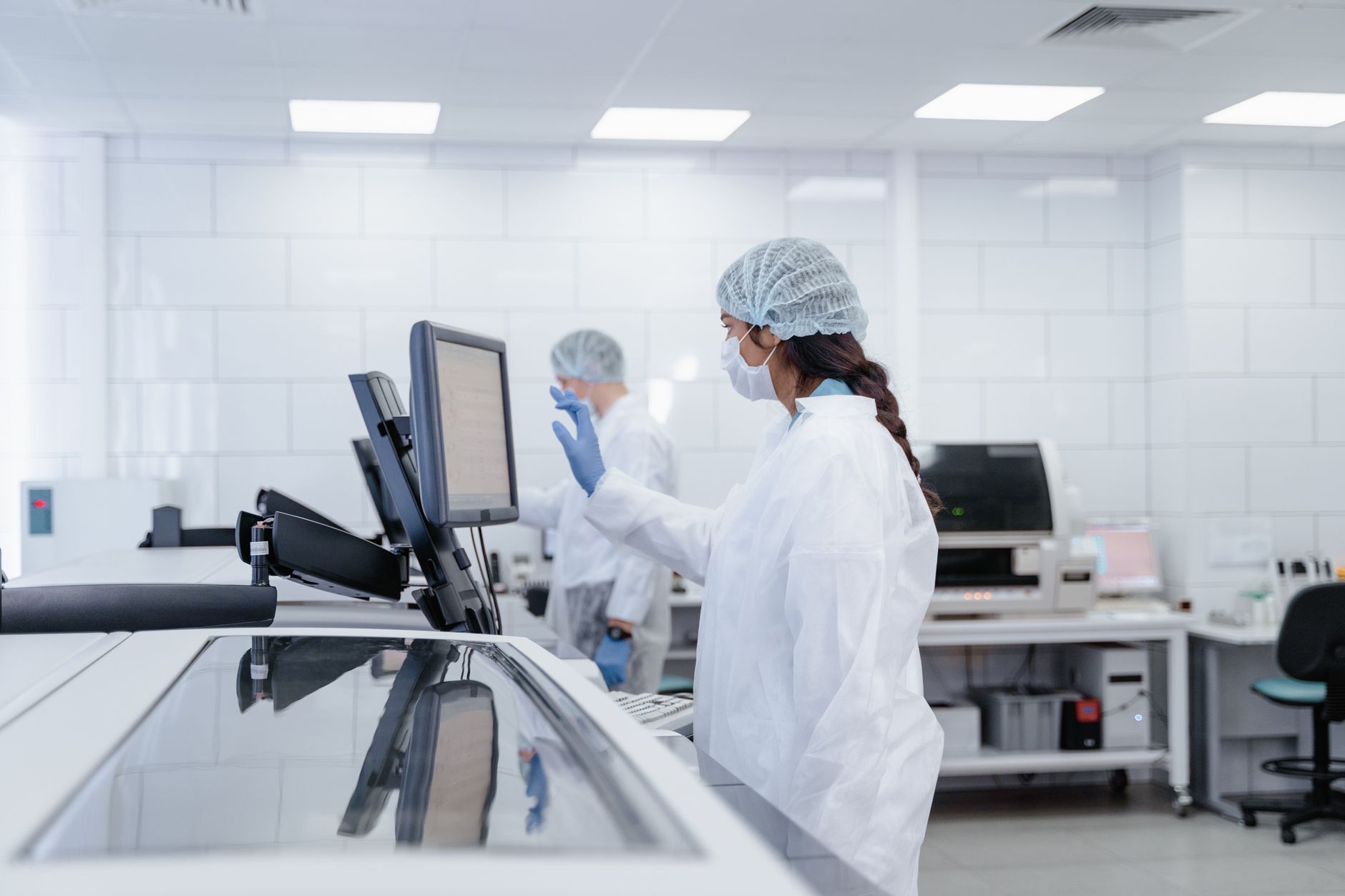 a researcher reviewing complex data visualizations on large dual monitors inside a clinical informatics lab, with rows of medical reference binders visible on shelves in the background