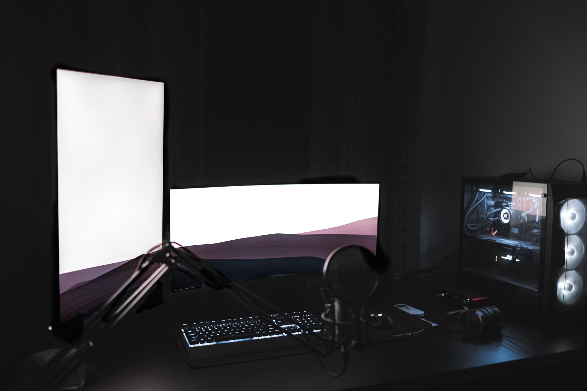 a dual-monitor streaming setup with overlay graphics visible on screen, RGB keyboard and capture card on a desk in a dim room