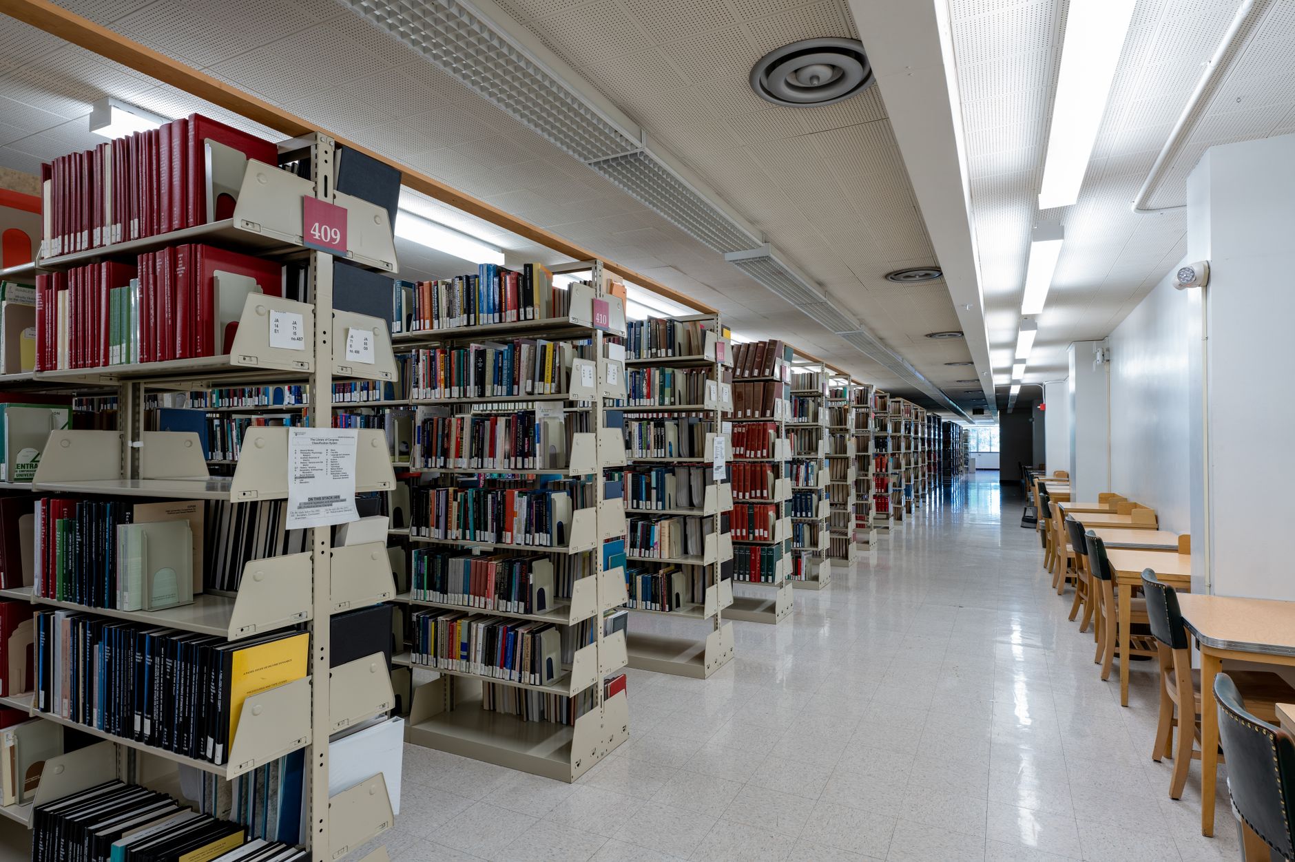 rows of physical research binders and printed journal articles stacked on metal shelving units inside a university archive room, with a single monitor glowing in the background
