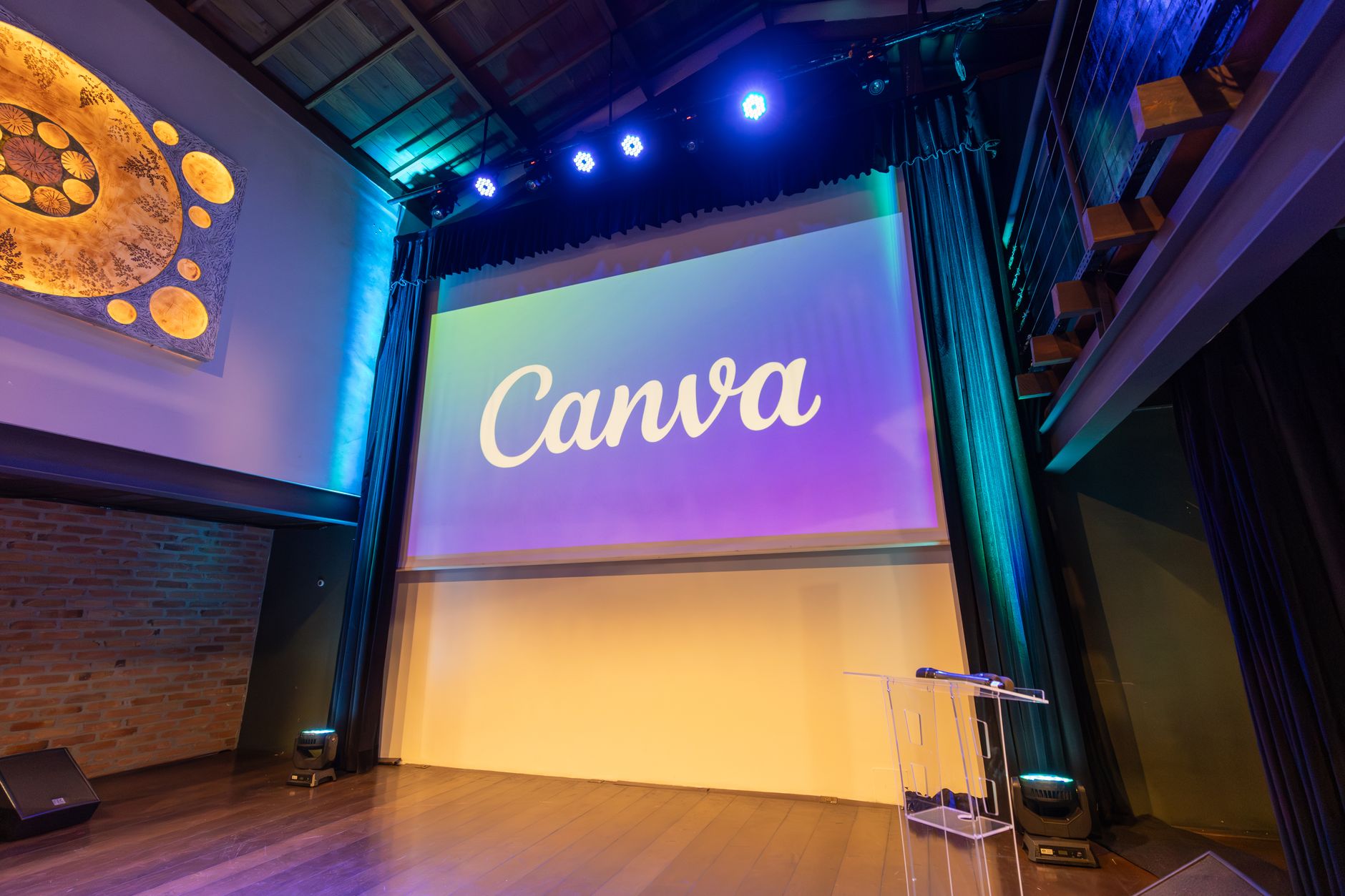 a wide-angle view of a modern convention hall interior with rows of standing-height tables, laptops open, and a large projection screen displaying a data visualization at the far end