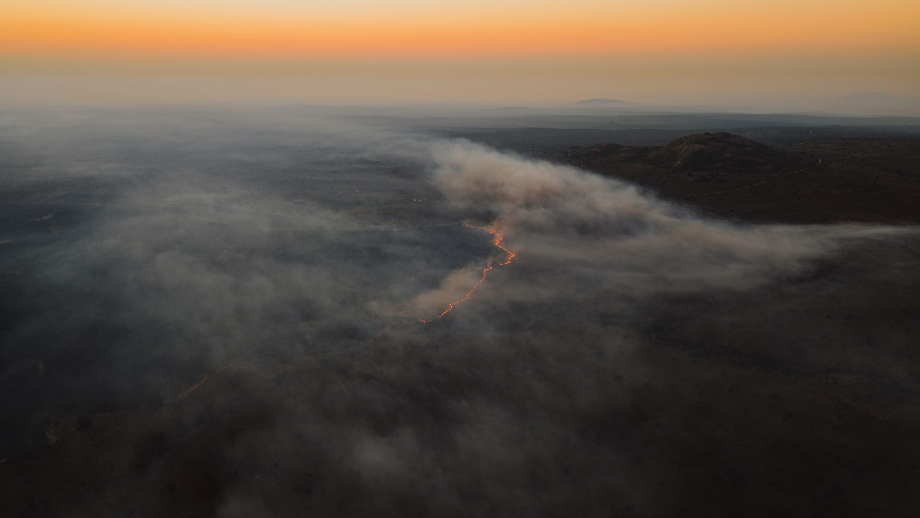 aerial view of a charred hillside landscape with burned structures visible among blackened trees, smoke haze in the distance