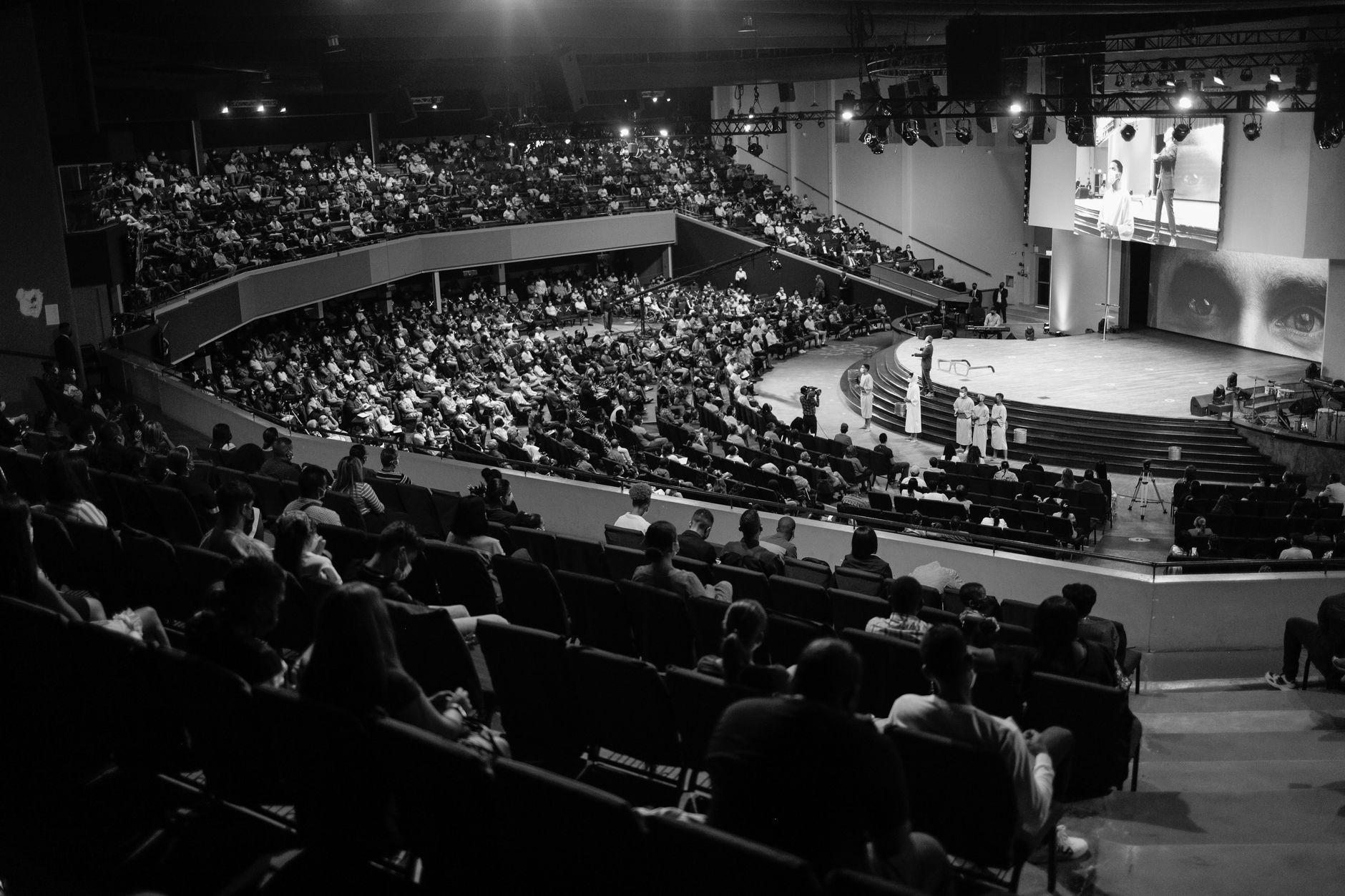 a cinematic wide shot of a darkened convention hall stage with a massive curved LED screen displaying a spacecraft in deep space, audience silhouettes visible in the foreground