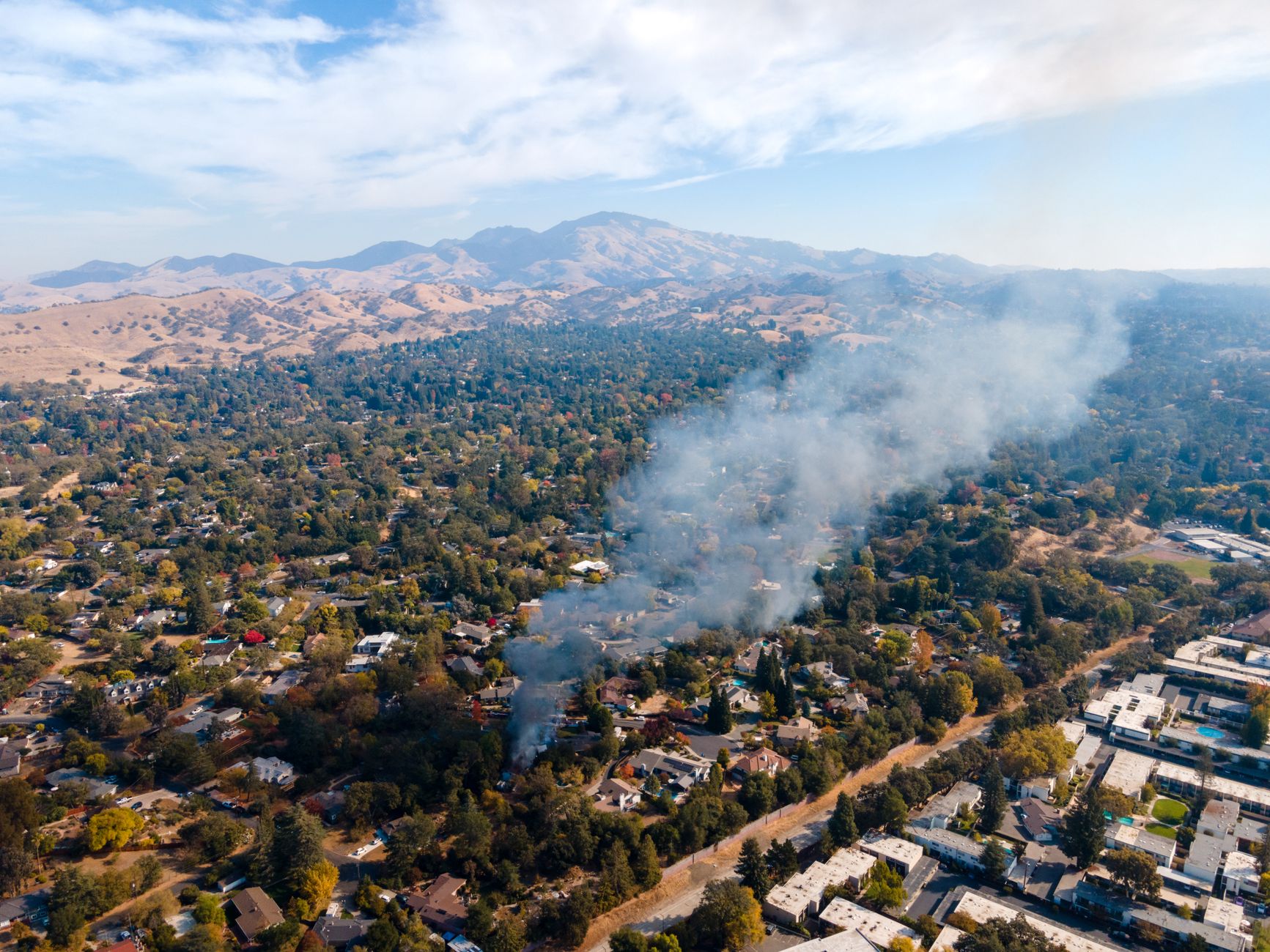 aerial view of a hillside neighborhood showing fire-scorched lots alongside standing homes, with ash-covered streets and power line corridors visible