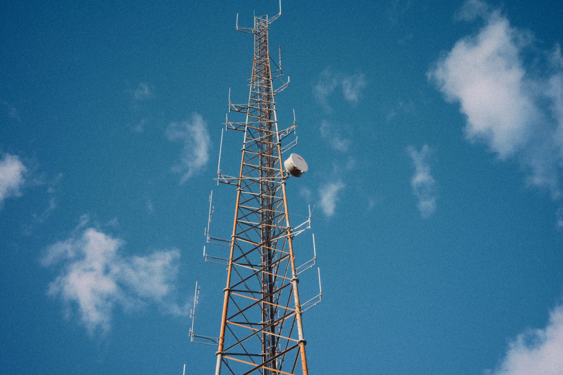 exterior of a large broadcast transmission tower against a cloudy sky, with satellite dishes mounted on an industrial rooftop visible below