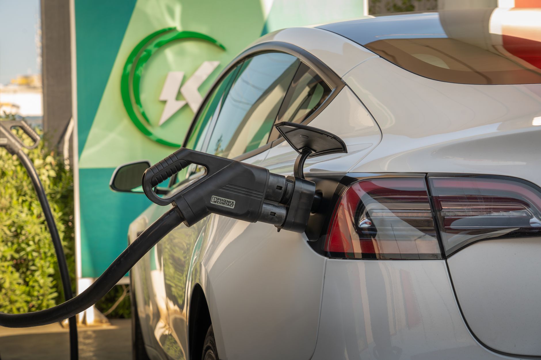 an electric vehicle charging station grid at a highway rest stop, multiple vehicles plugged in under a canopy structure at dusk with distant hills in the background