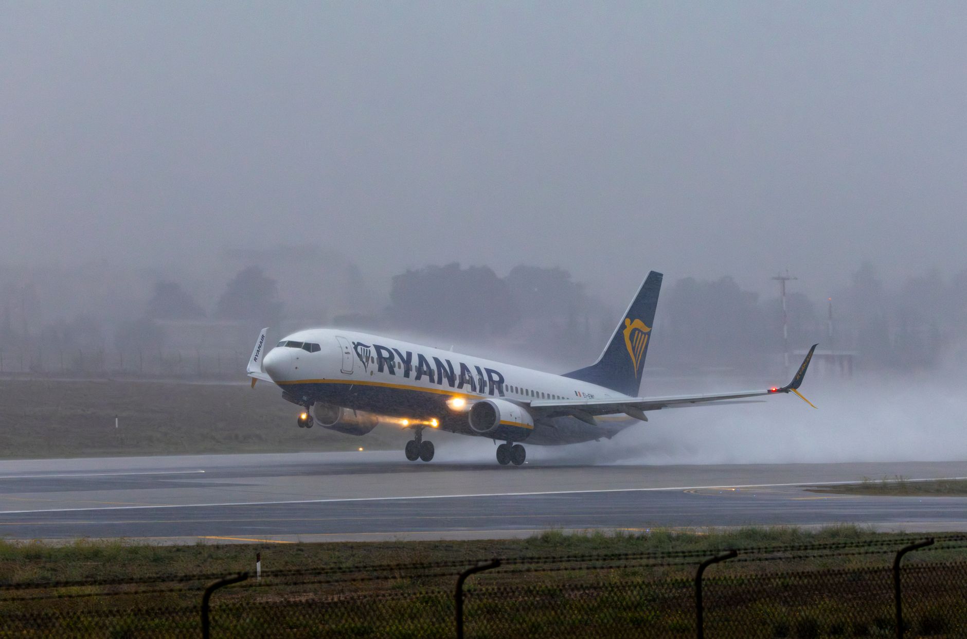 a large cargo aircraft on a rain-slicked airport tarmac at dusk, emergency vehicle lights visible in the far background near a runway