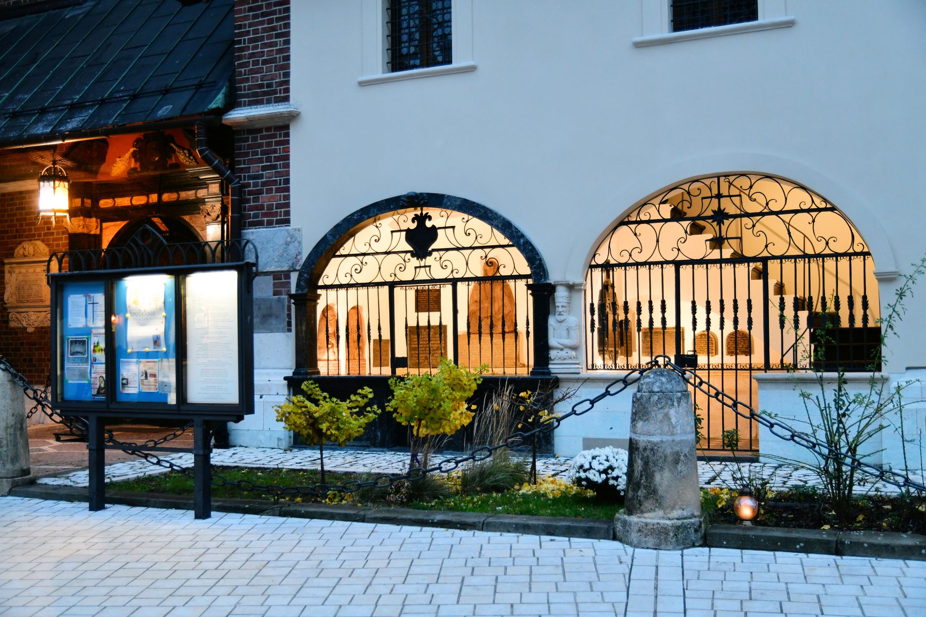 exterior view of the West Wing entrance at dusk, iron security fence in the foreground, warm interior lights visible through tall windows