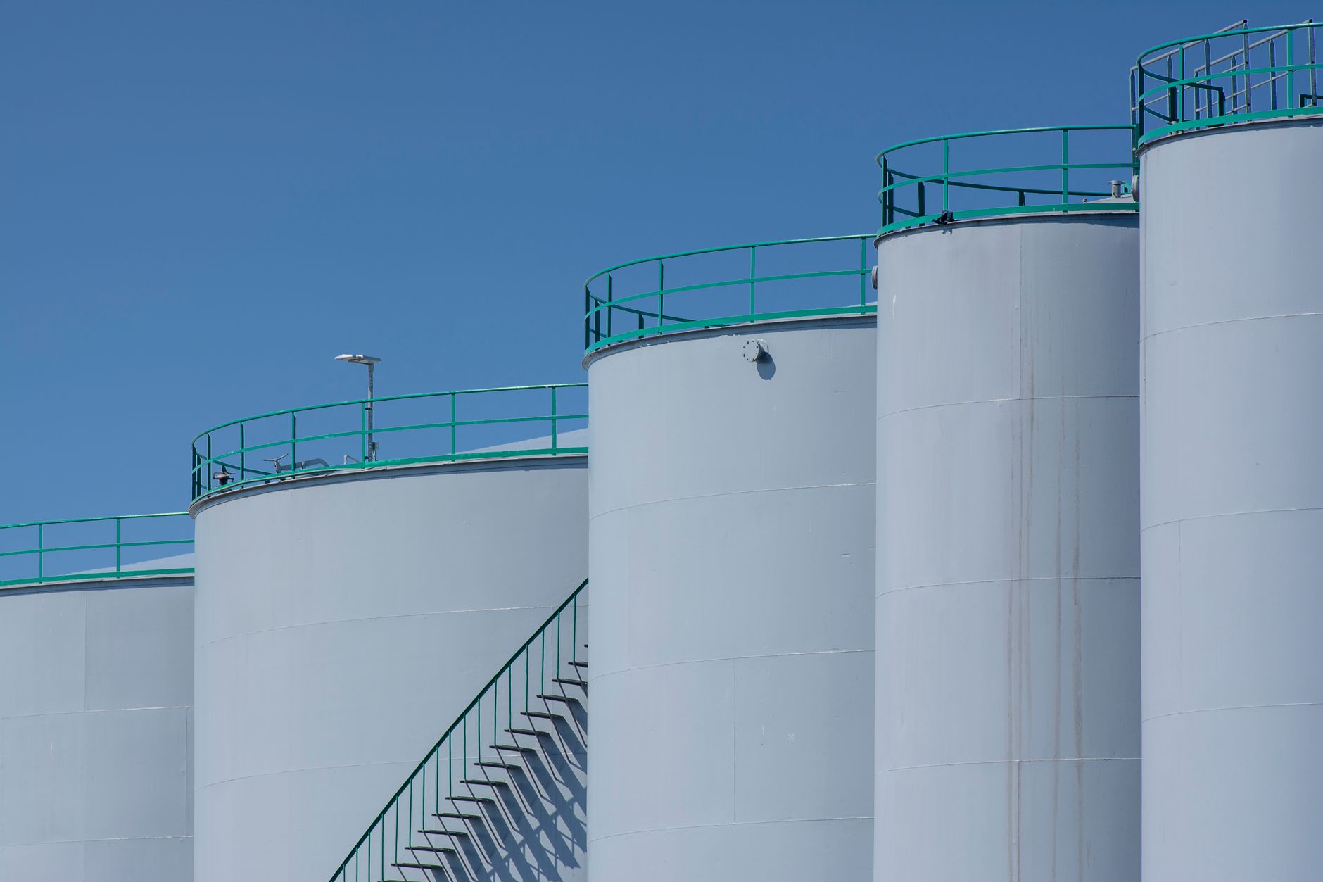 rows of large cylindrical crude oil storage tanks in a flat industrial facility, pipe infrastructure visible between units under an overcast sky