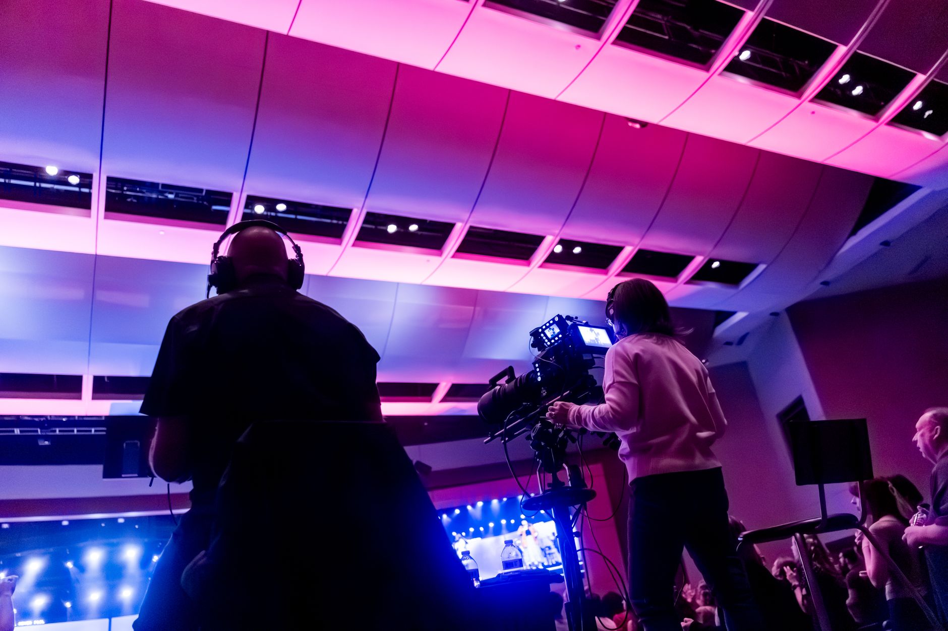 an empty late-night television studio stage with dramatic overhead lighting, a single desk and chair at center, camera equipment visible in the foreground