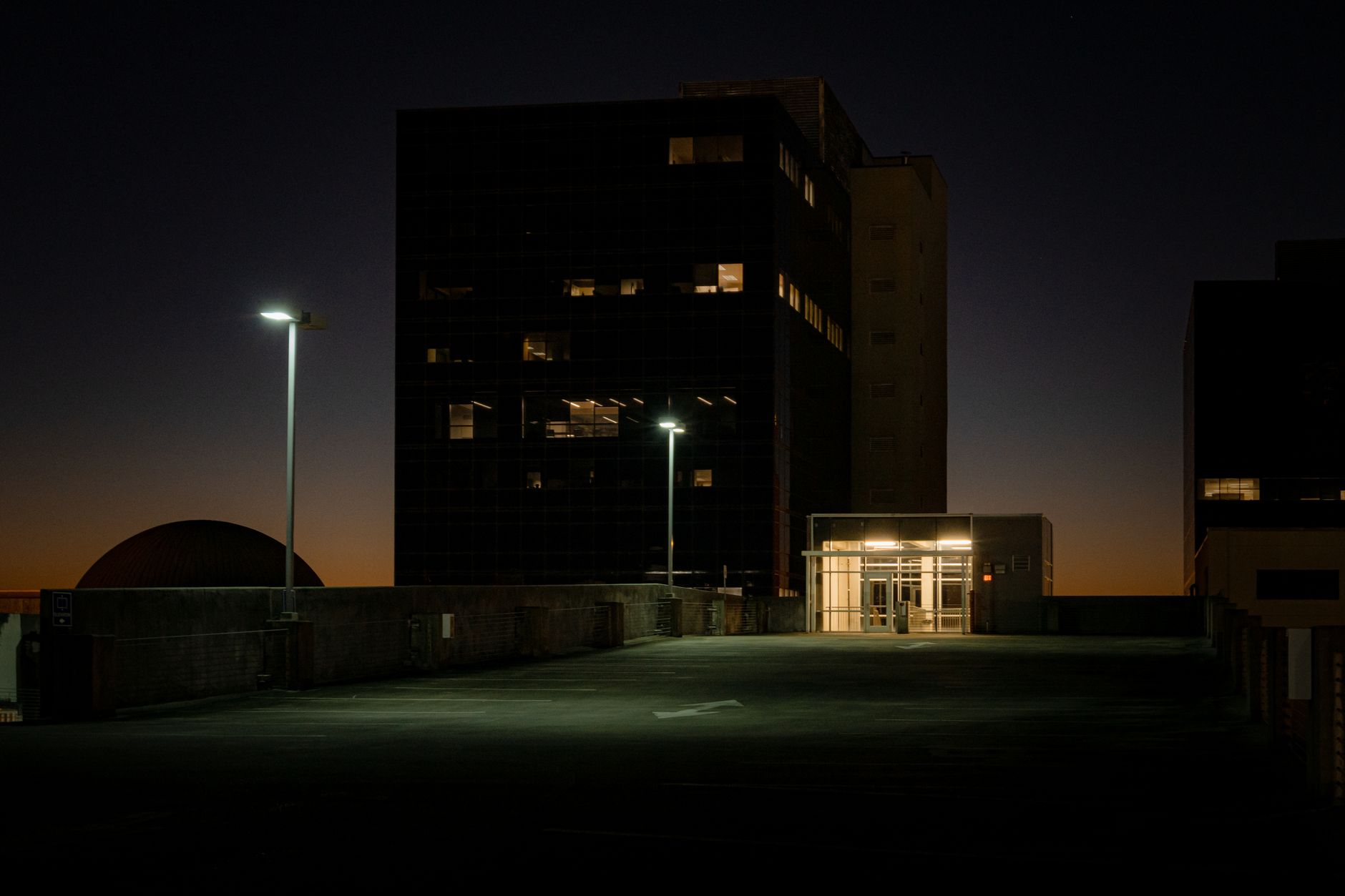 a wide-angle shot of a modern AI research campus at dusk, glass-walled offices lit from within, empty outdoor walkways visible between buildings