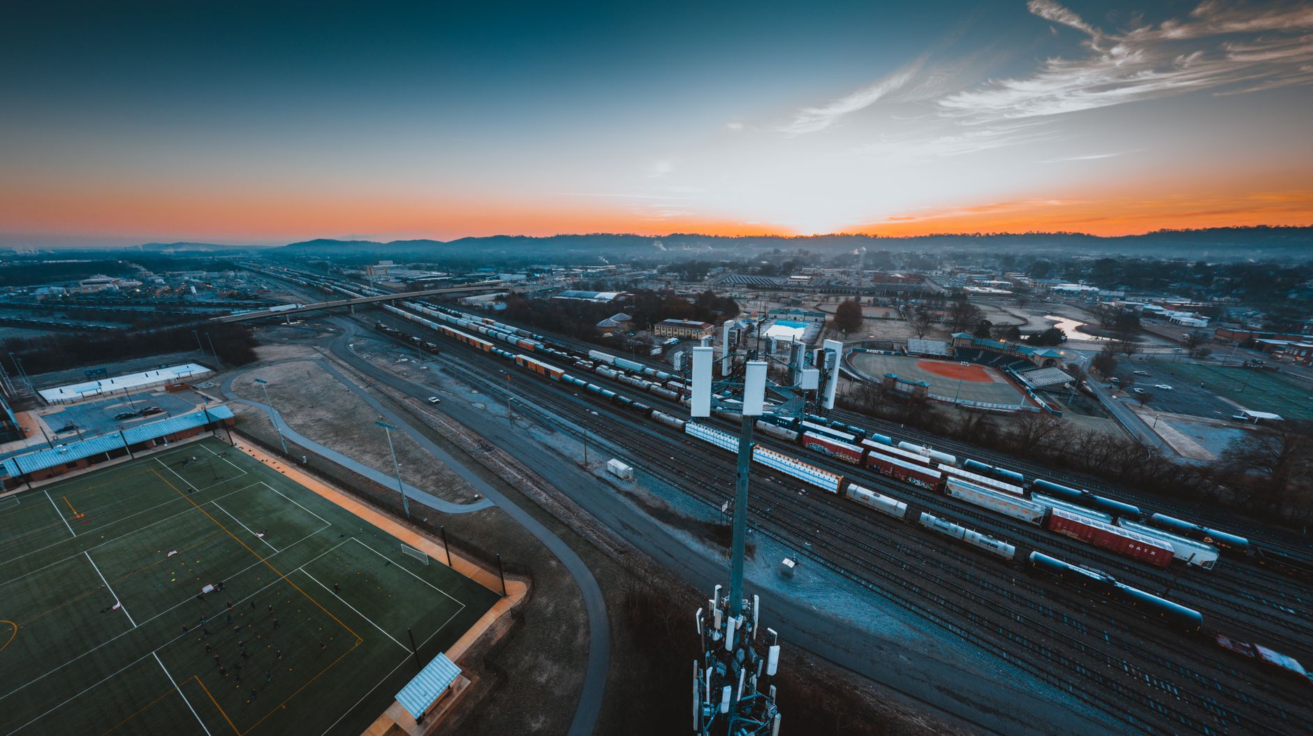 an aerial shot of a large corporate campus perimeter at dusk, with security camera poles and controlled-access gates visible along the boundary