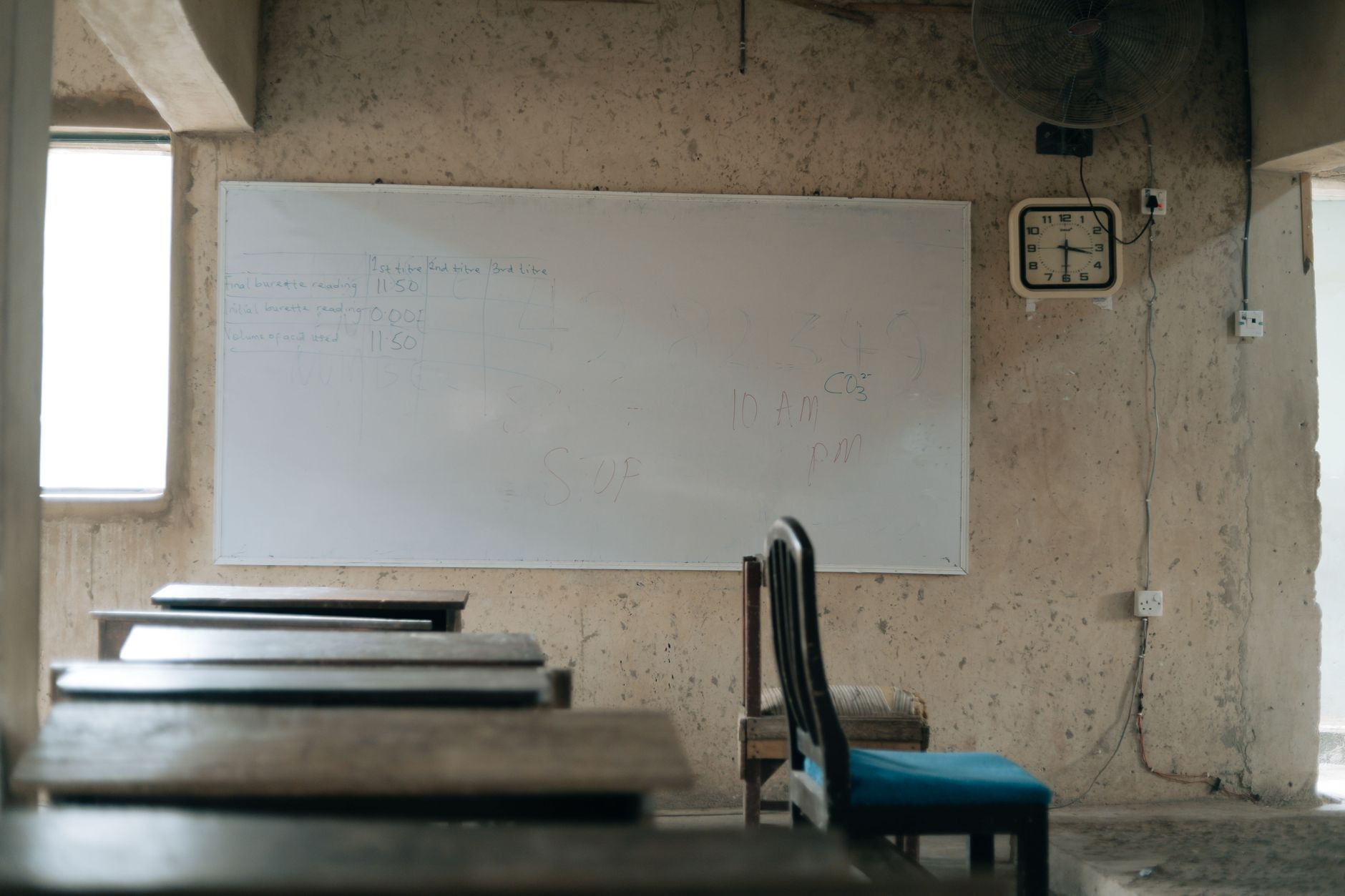 rows of empty wooden school desks inside a sunlit Israeli classroom, textbooks stacked at the front near a chalkboard covered in equations