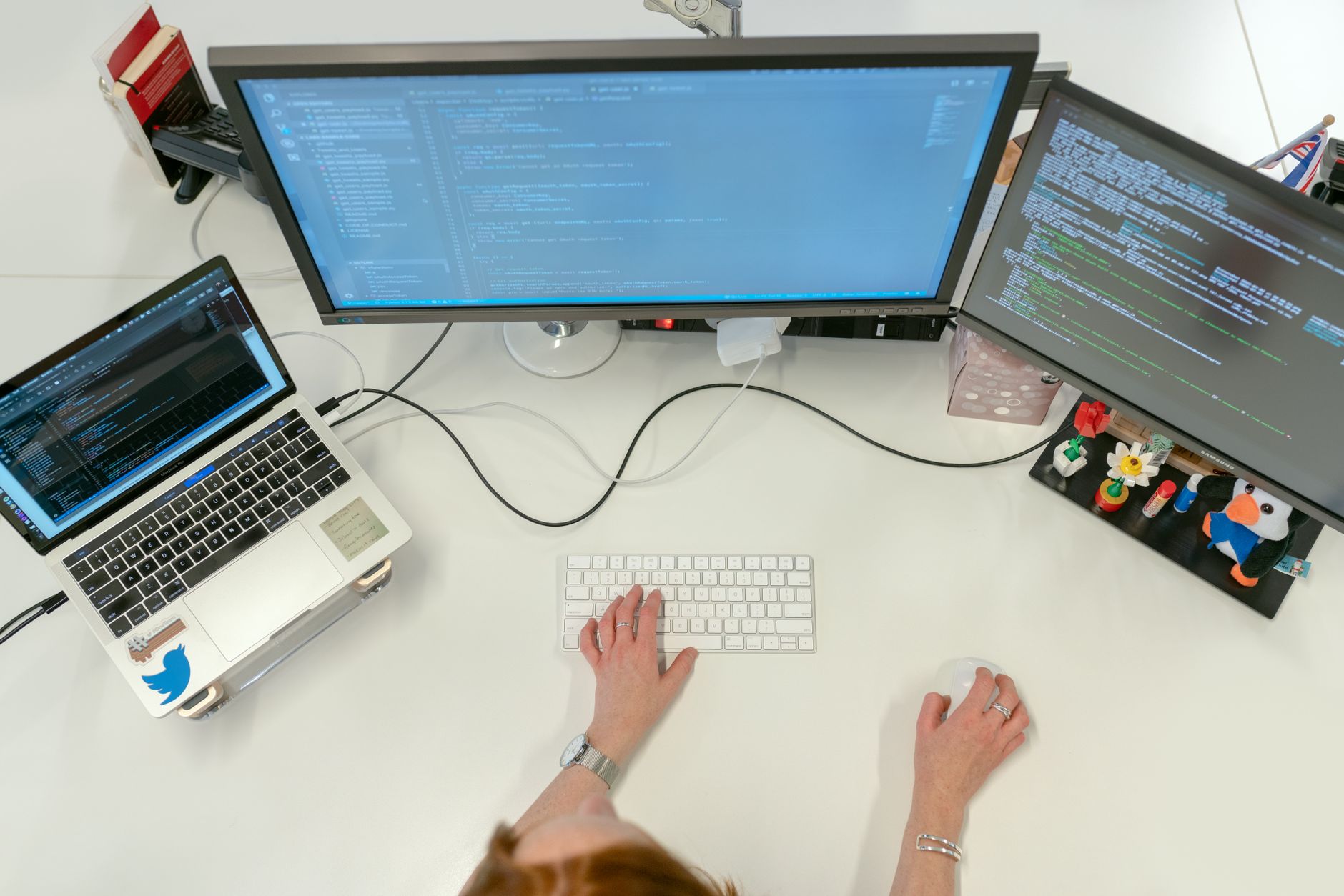 A wide shot of a busy technology hub co-working space with rows of standing desks, multiple monitors displaying code editors and analytics tools, natural light streaming through floor-to-ceiling windows