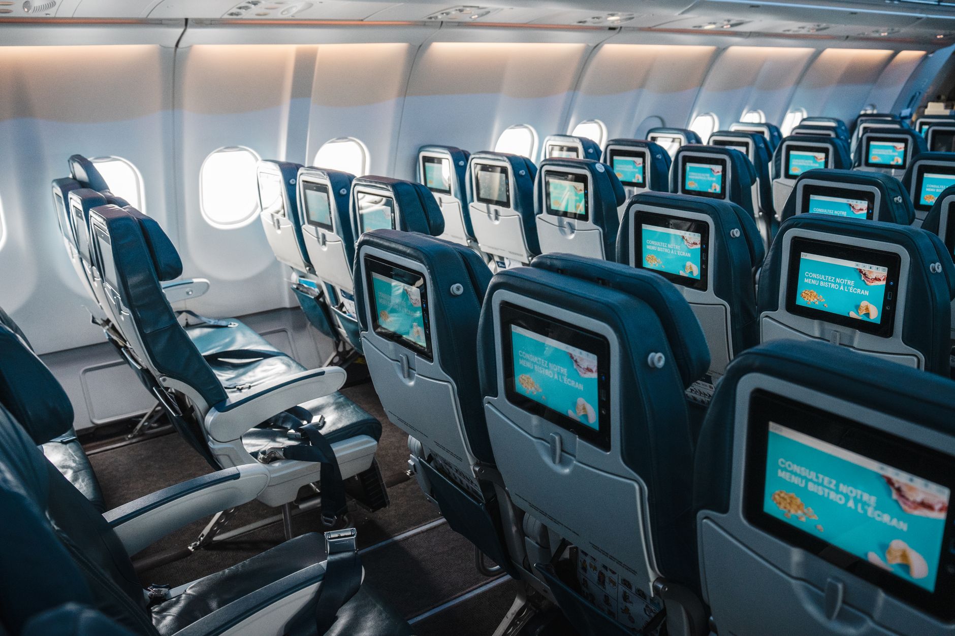 rows of economy-class airplane seats inside a wide-body aircraft cabin, lit by overhead lighting with window shades partially open
