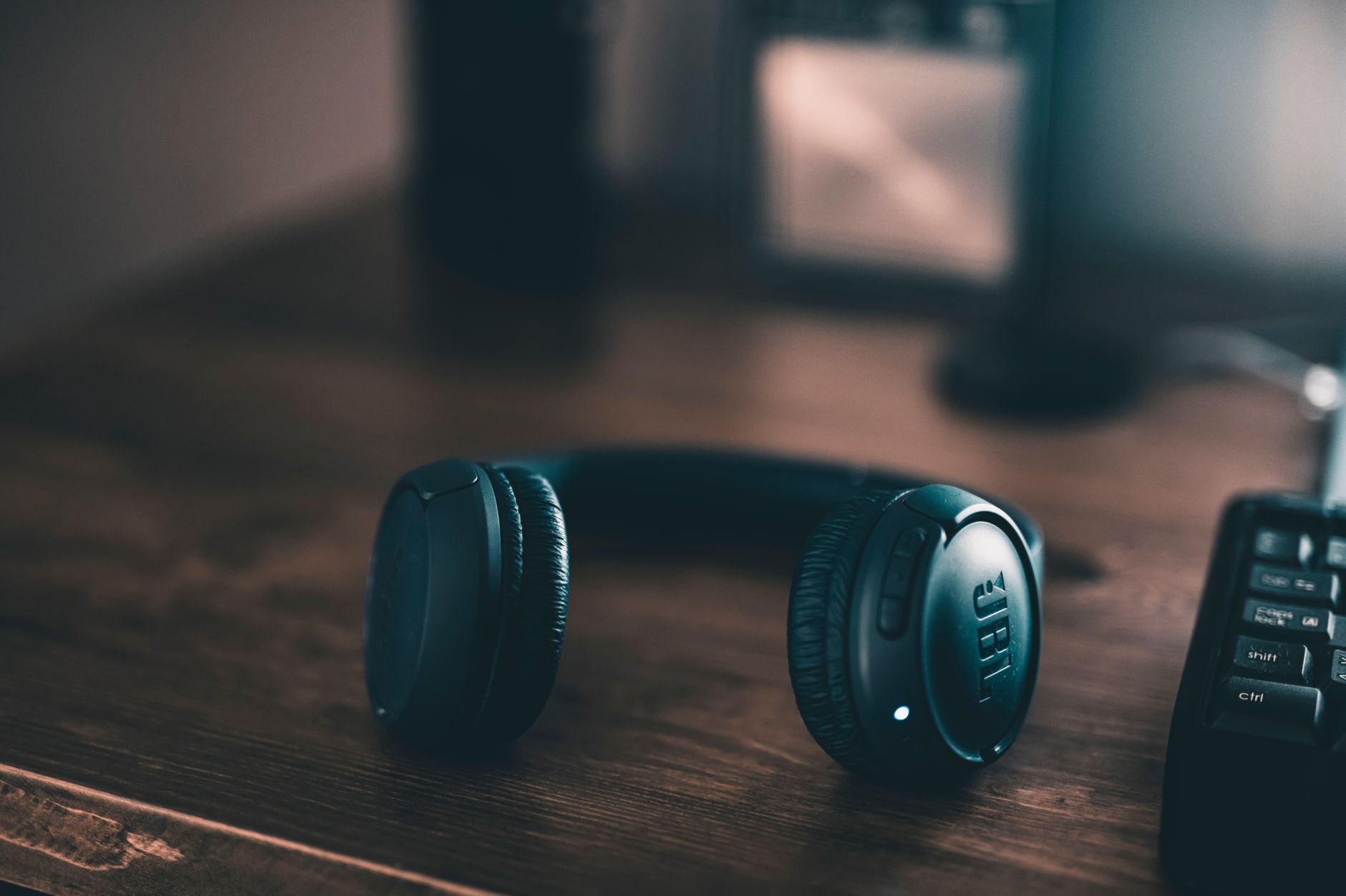 A close-up of a black wireless gaming headset resting on a desk next to a gaming controller, with soft ambient lighting highlighting the ear cup design and headband padding