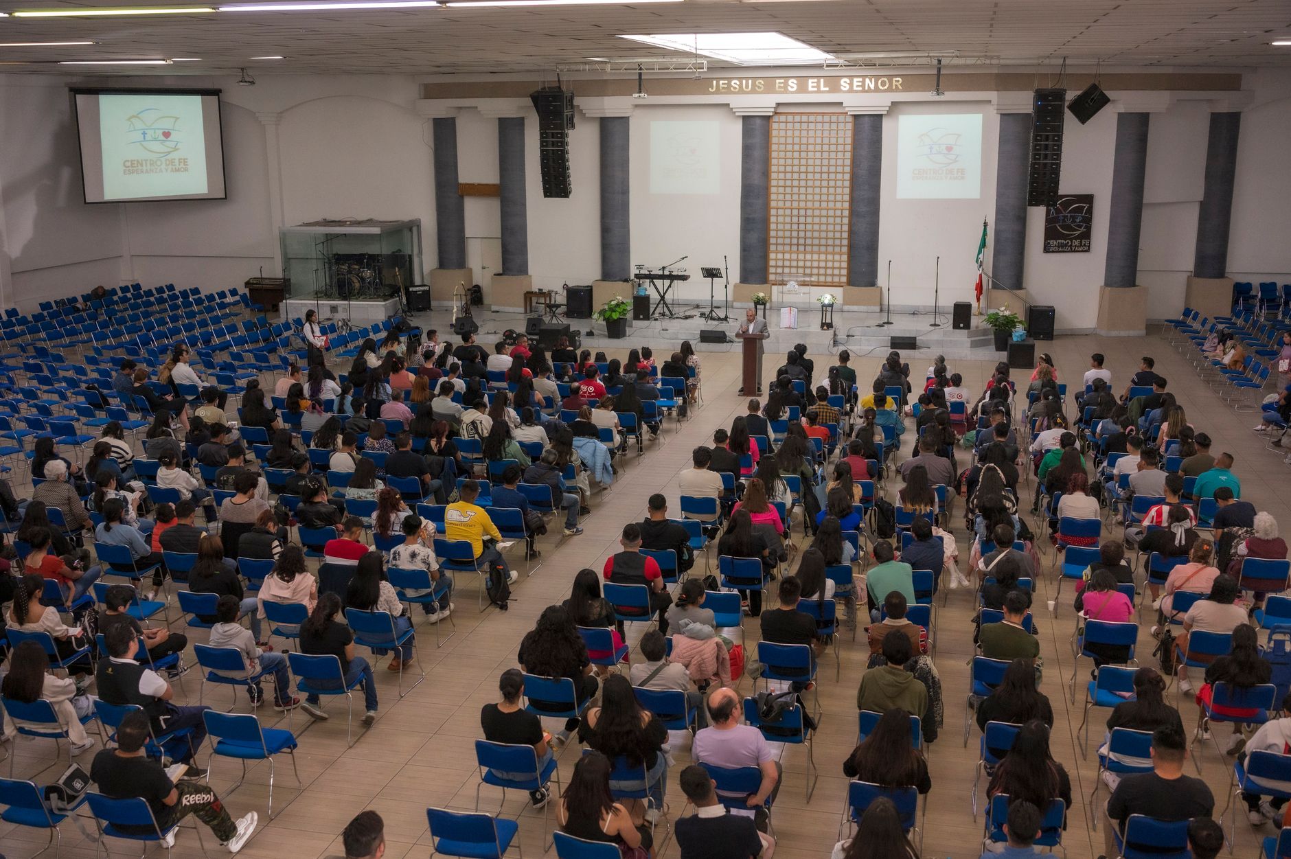 a large lecture hall with rows of empty wooden seats and a projection screen at the front showing a split-screen of a traditional slide deck beside a vertical video interface