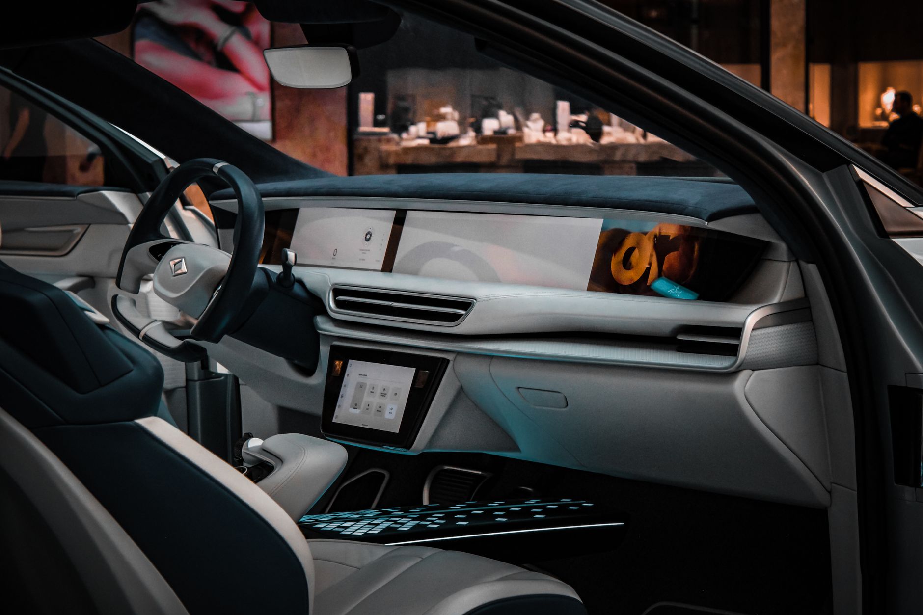 a sleek, pedal-free two-seat autonomous vehicle parked at a curbside rideshare pickup zone in an urban setting, viewed from the front quarter angle showing the absence of a steering column through the windshield