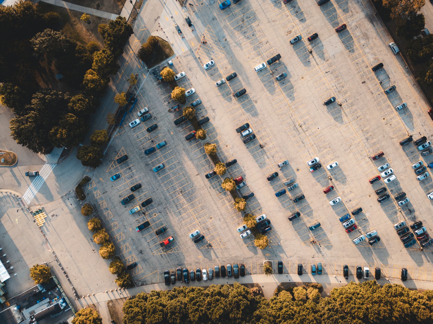 a row of compact autonomous vehicles queued at a charging station in an open urban parking facility, late afternoon light casting long shadows across the pavement