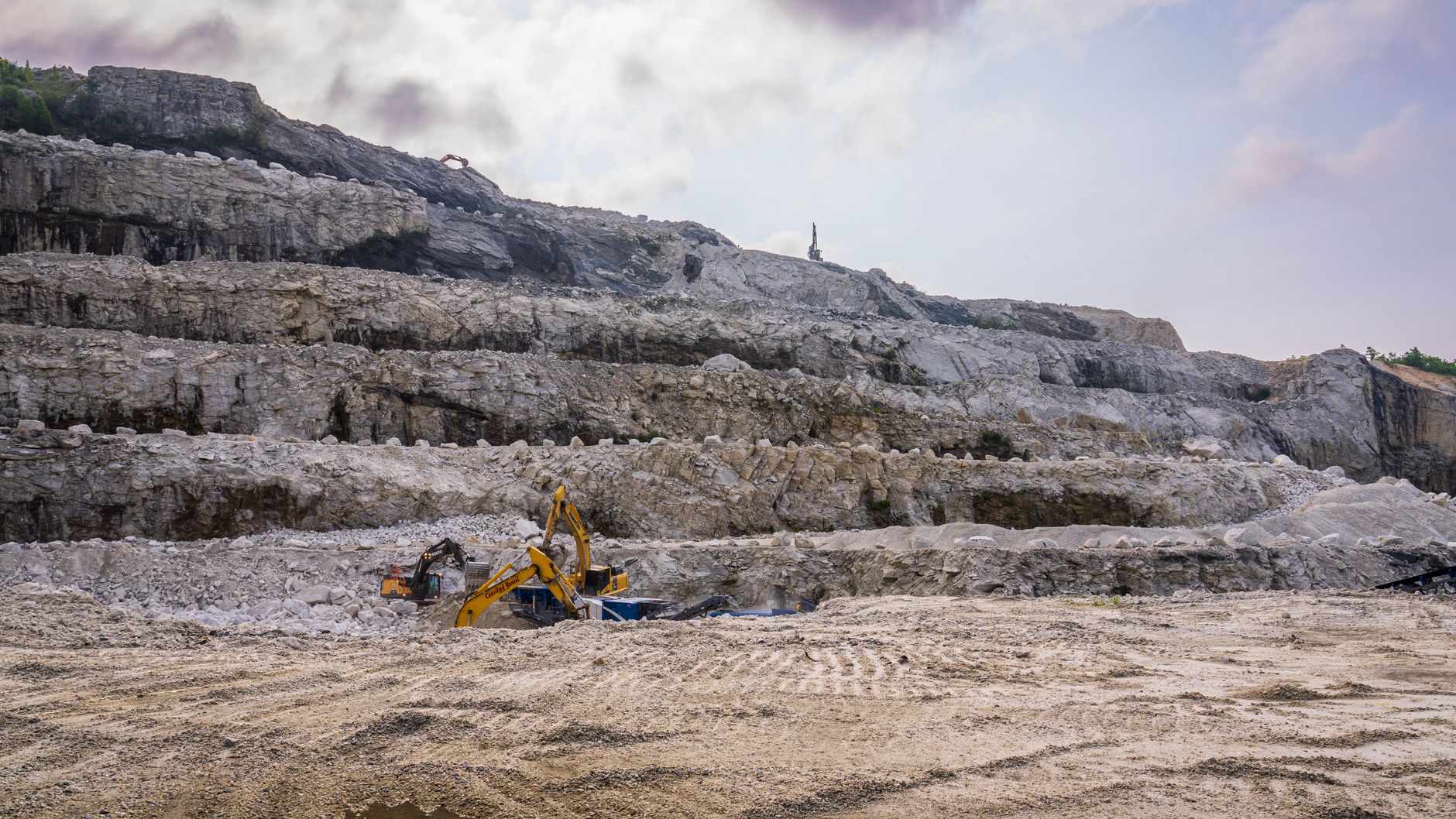Open-pit mining operations at a large rare-earth mineral extraction site, with heavy machinery working terraced rock faces under a pale sky