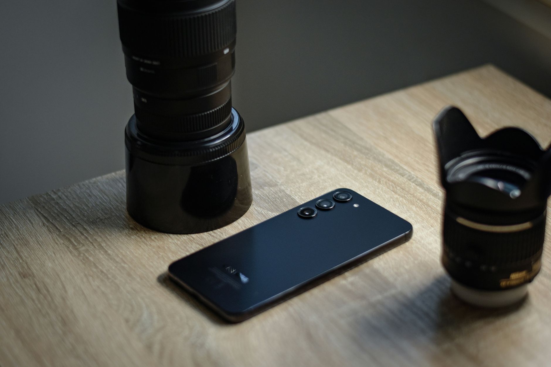 a close-up of a matte-finish smartphone resting on a wooden desk, its camera module visible with multiple lenses catching soft ambient light
