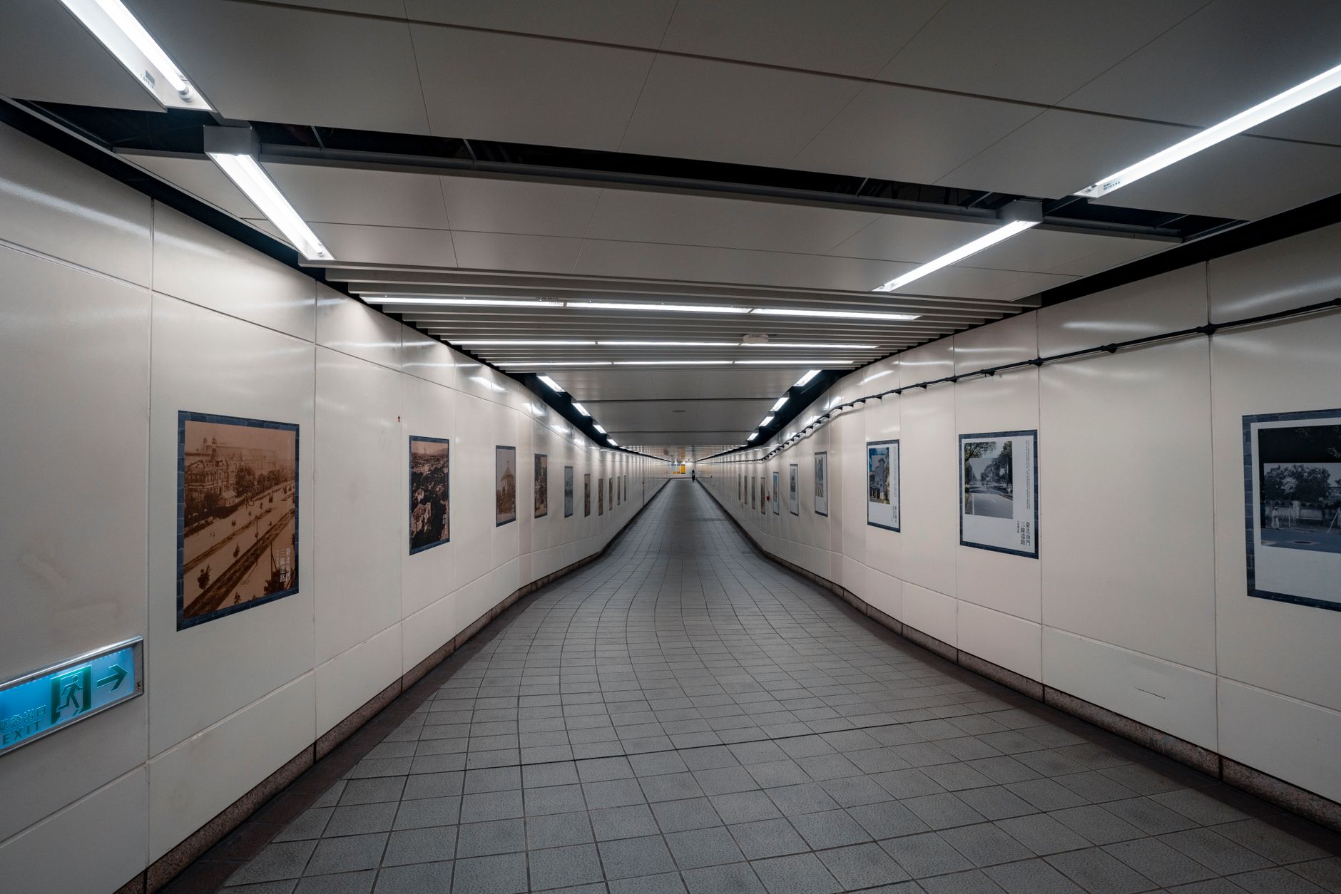 a narrow institutional corridor with a wall-mounted video kiosk terminal, fluorescent lighting overhead, metal benches visible along the wall
