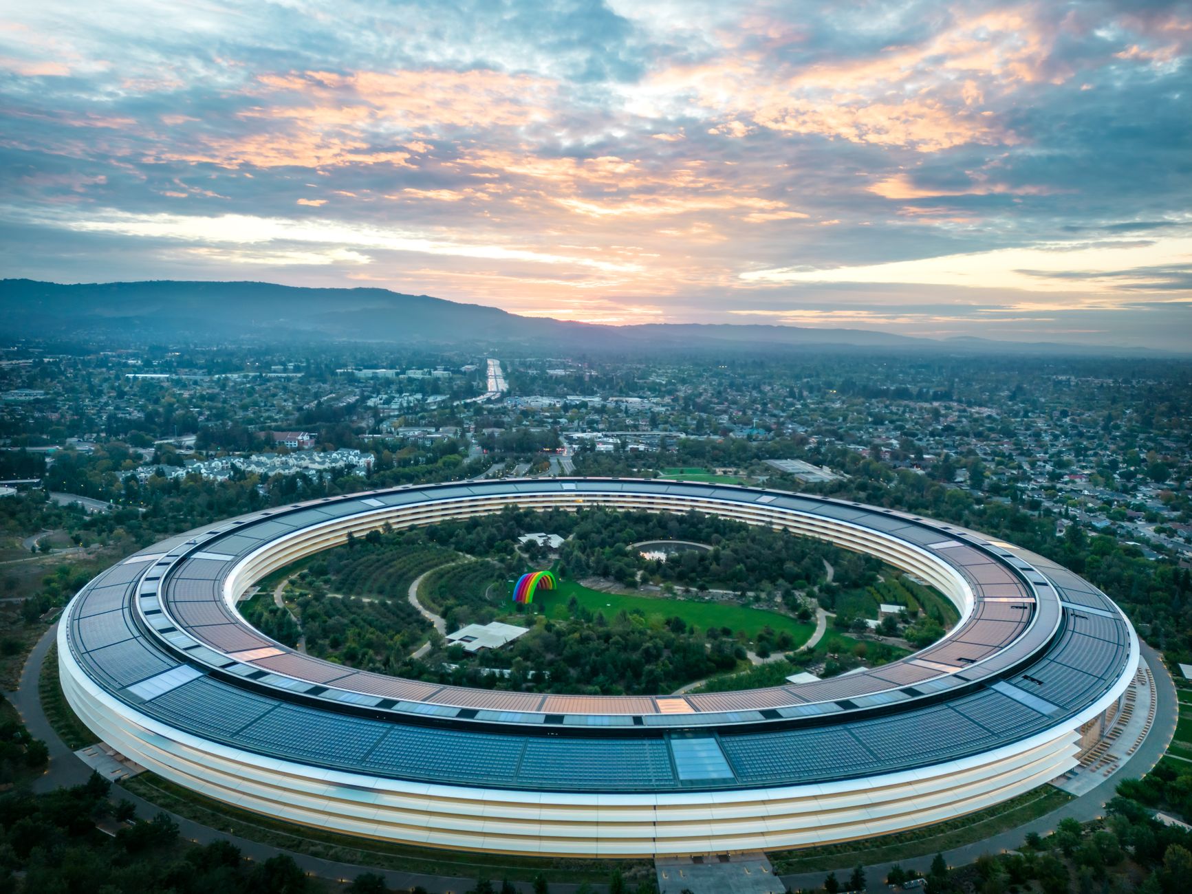 aerial view of a modern urban technology campus at dusk, glass buildings illuminated with interior lights, surrounded by manicured green spaces