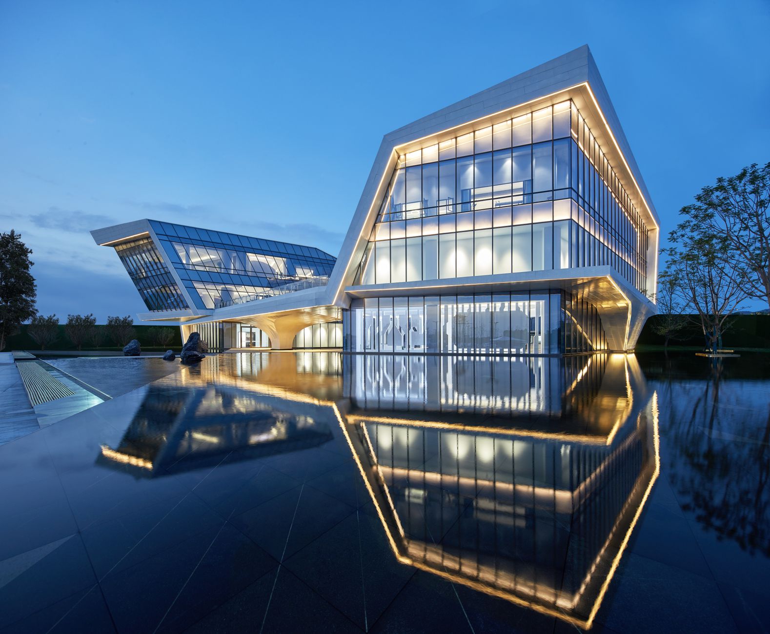 A wide-angle view of a modern innovation campus at dusk, glass-fronted research buildings lit from within, surrounding a central open plaza with sparse trees