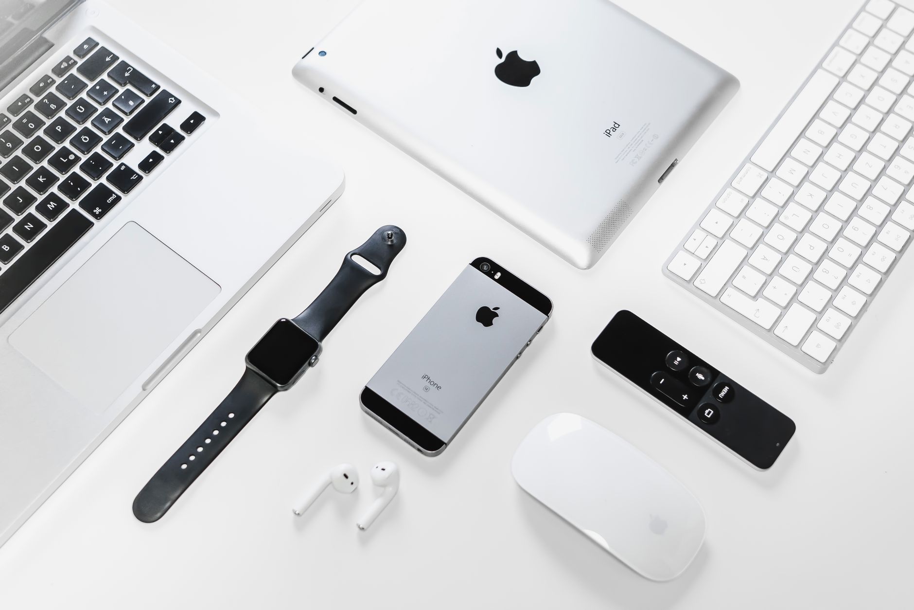 a flat-lay of consumer electronics including a robot vacuum, wireless earbuds, and a smart speaker arranged on a light grey surface