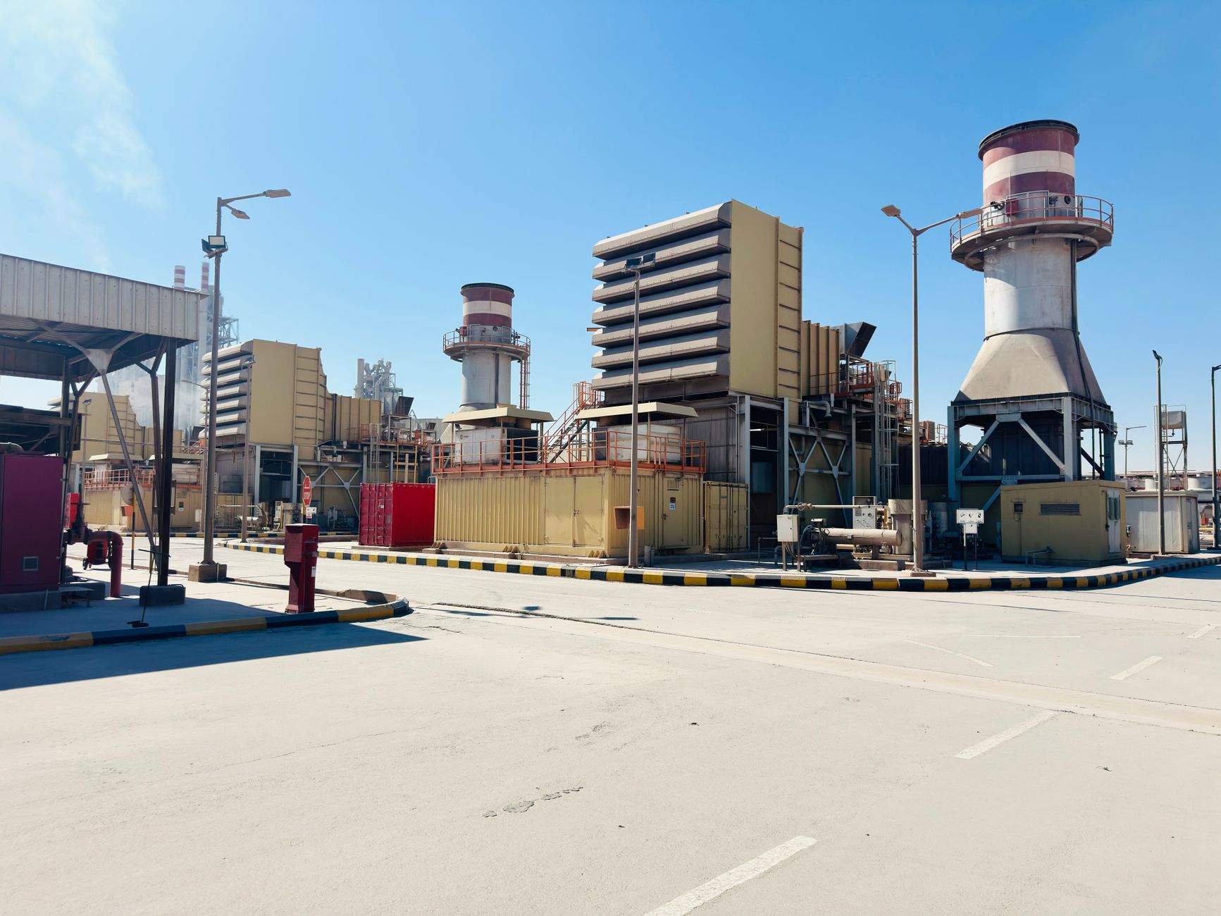 a small distributed power generator unit mounted on a concrete pad in an industrial yard, photographed at midday with industrial equipment visible in the background