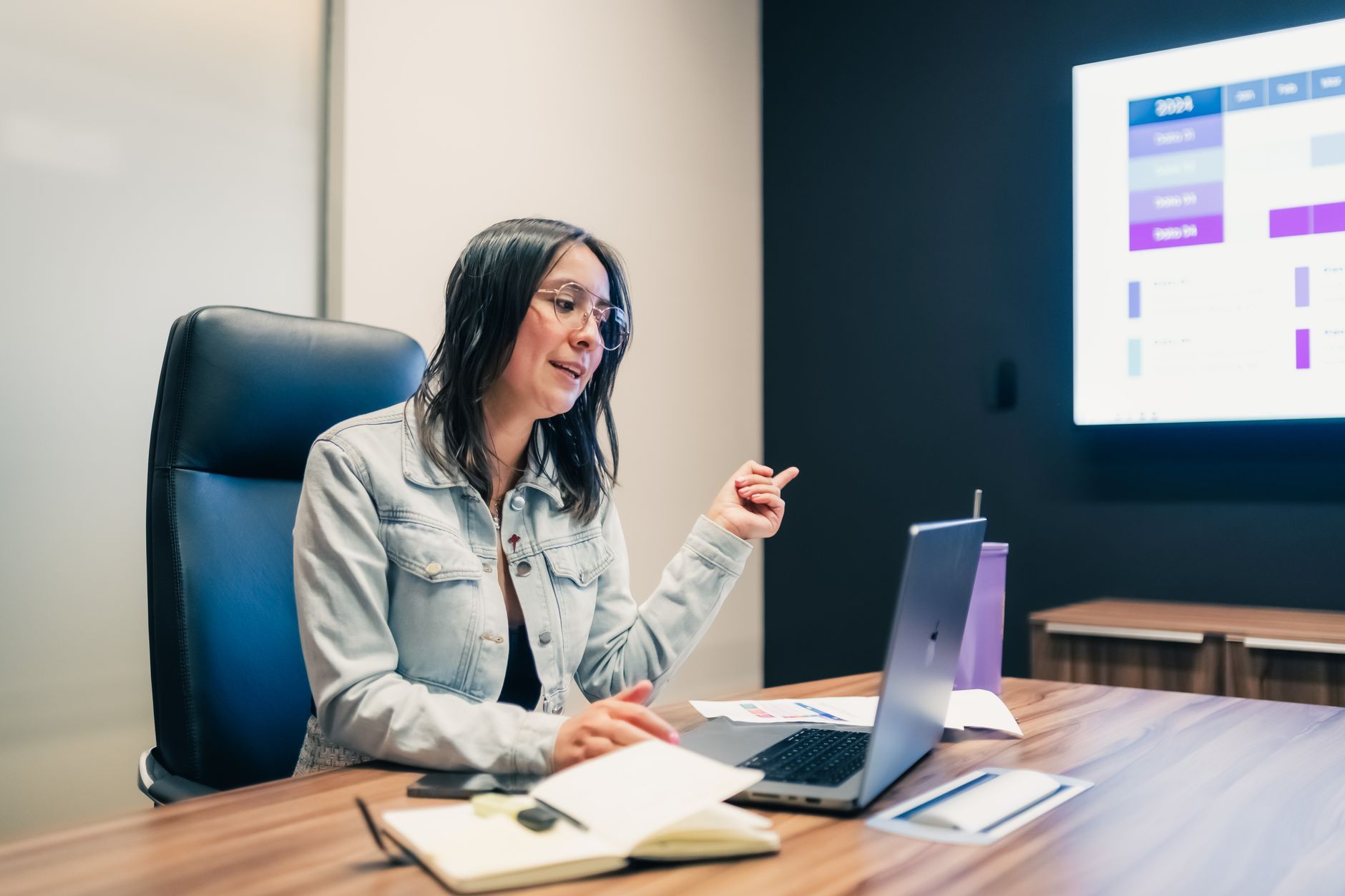 A softly lit office conference room with a large wall-mounted screen displaying abstract neural network visualizations, an empty round table with open laptops and water glasses in the foreground